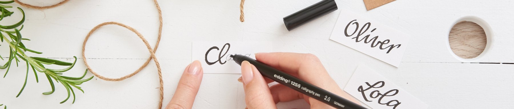 Die Hand einer Person hält einen schwarzen Stift und kreiert elegante Kalligraphie auf einer weißen Tischkarte, mit weiteren fertigen Karten und dekorativem Grün, das kunstvoll auf einer weißen Holzoberfläche verstreut ist.