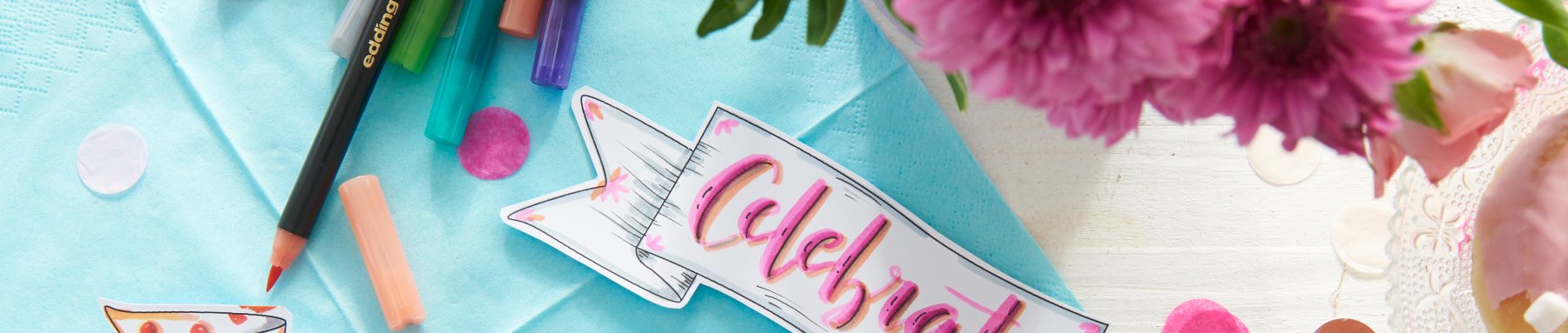 Ein lebendiger, kreativer Arbeitsbereich mit Malutensilien, einem „Feier“-Banner und frischen rosa Blumen, die eine festliche und inspirierende Atmosphäre symbolisieren.