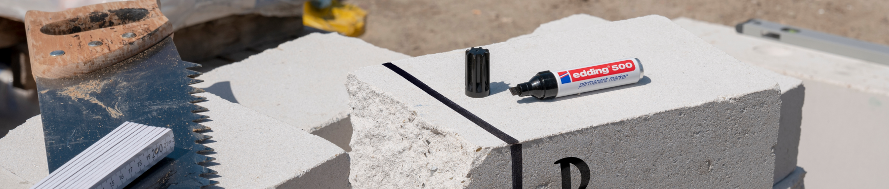 Eine Bauszene mit einer Nahaufnahme von Betonblöcken mit einer Tube Klebstoff mit der Aufschrift „tecbond 500“, daneben ein Sägeblatt und ein schmutziger Arbeitsstiefel, was auf laufende Bau- oder Reparaturarbeiten hinweist.
