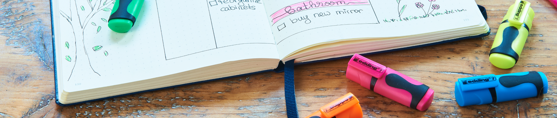 Verschiedene bunte Textmarker verstreut um ein offenes Notizbuch mit handschriftlichen Notizen auf einer Holzoberfläche.