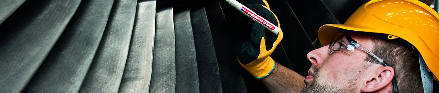 Ein konzentrierter Arbeiter mit gelbem Schutzhelm und Schutzbrille inspiziert schwere Maschinen mit einer Taschenlampe.