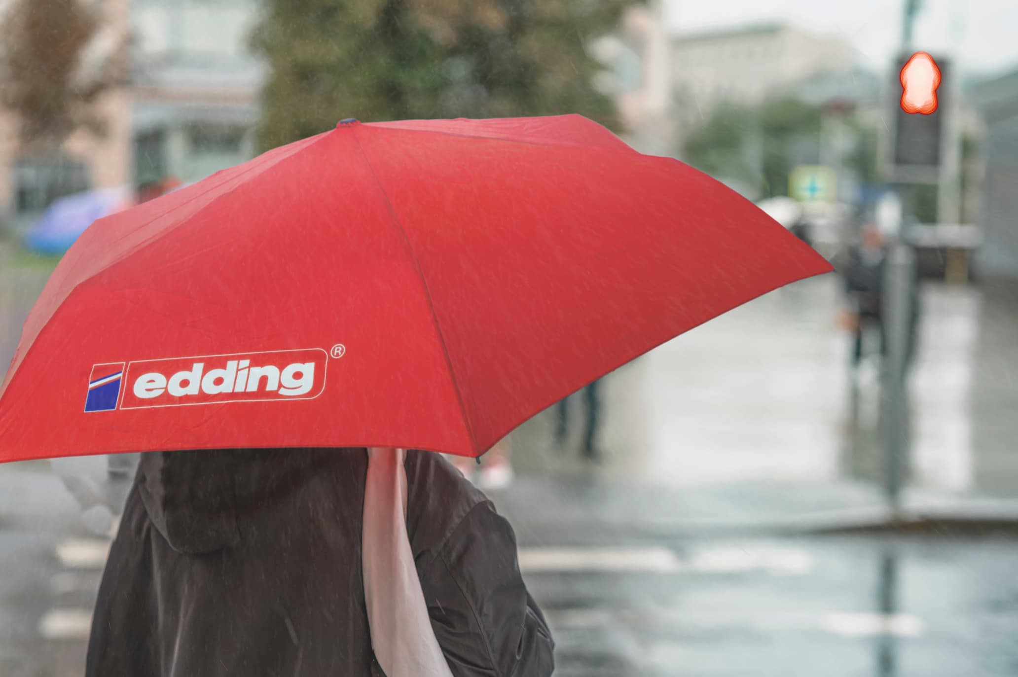 A person with a large edding Taschenschirm crafted from recycelte Kunststoffe strolls through the rain on a city street. In the backdrop are blurred buildings, a pedestrian crossing, and a red traffic light.