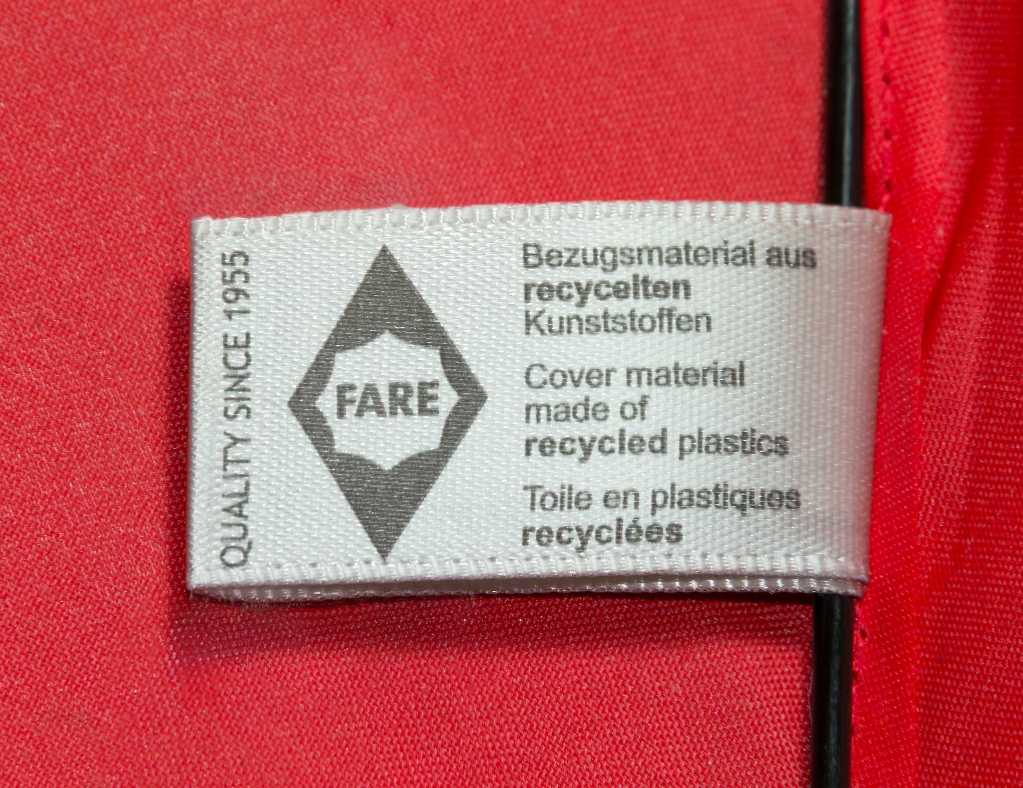 A clothing label on red fabric reads: "QUALITY SINCE 1955" and "Cover material made of recycled plastics" in many languages. Next to it is a hexagonal logo with "edding," indicating it's from the sustainable edding Taschenschirm collection.