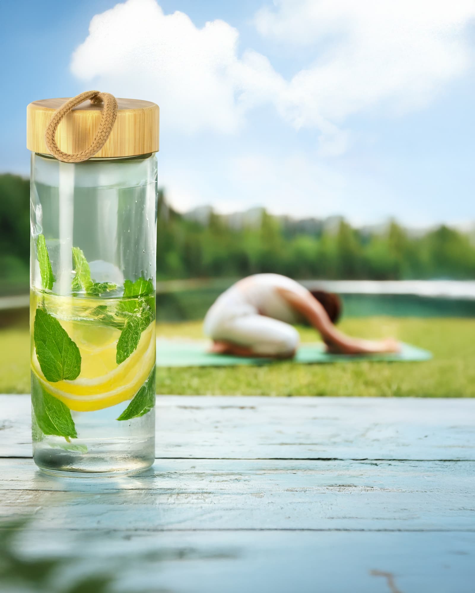 Eine edding Glasflasche 650 ml gefüllt mit Zitronen-Minze-Wasser steht auf einer Holzoberfläche. Im unscharfen Hintergrund praktiziert jemand Yoga auf einer grünen Matte, umgeben von Gras und Bäumen unter einem blauen Himmel.