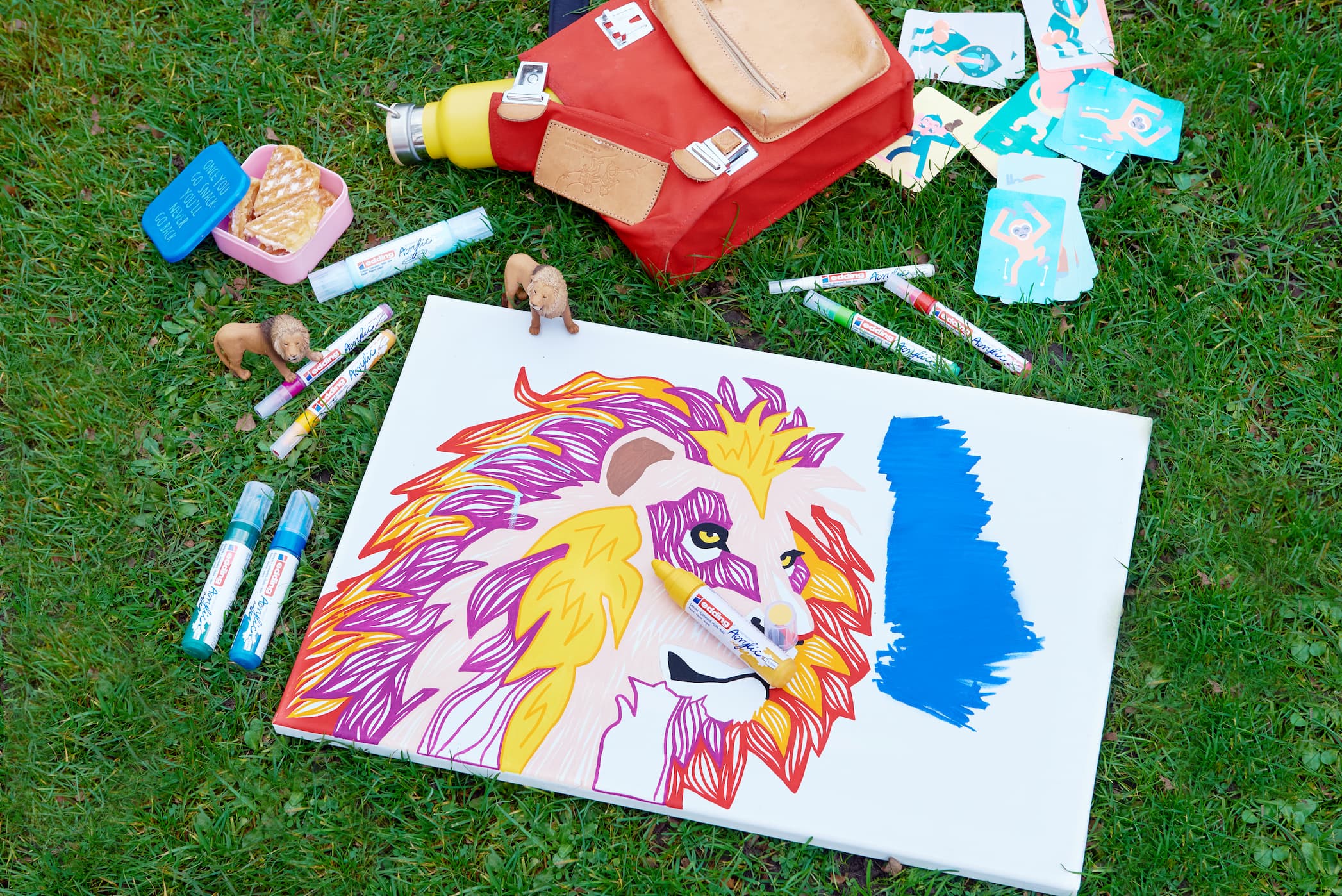 Im Gras verstreute Künstlerutensilien rund um ein unvollendetes, lebendiges Gemälde eines Löwengesichts, gemalt mit hochpigmentierter Tinte und einem edding 5000 Acrylmarker breit.