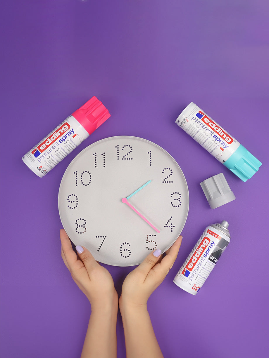 Eine Person mit manikürten Nägeln hält eine DIY Eule: Upcycling-Idee Wanduhr im Betonlook vor einem violetten Hintergrund. Rund um die Uhr stehen drei Dosen edding SHOP Permanentspray in Pink, Türkis und Grau, daneben liegen die Deckel – perfekt für eine kreative DIY Eule Upcycling-Idee.
