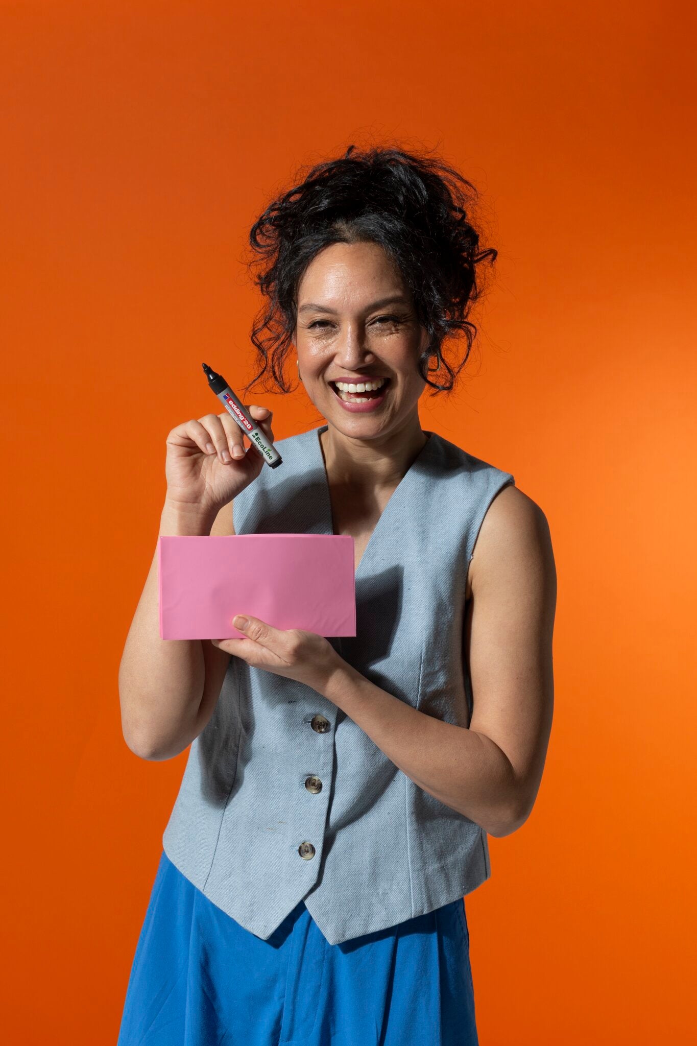 Eine lächelnde Frau steht vor einem orangefarbenen Hintergrund, hält eine PLAYROOM ESSENTIALS Kreativbox Magic-Chart von Playroom und einen schwarzen Marker in der Hand und trägt eine hellblaue Weste und eine blaue Hose.