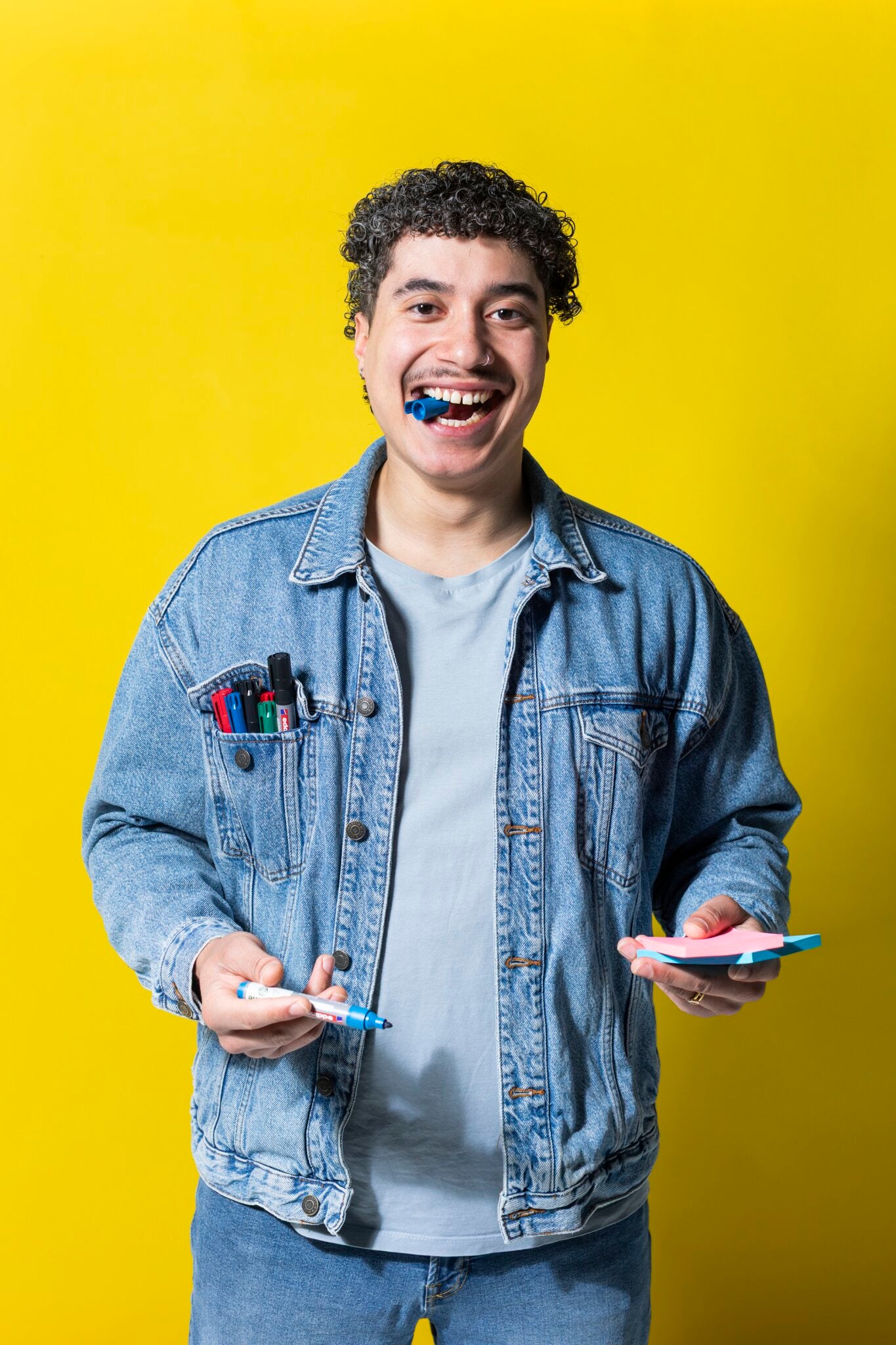 Eine lächelnde Person in Jeansjacke hält die PLAYROOM ESSENTIALS Kreativbox Workshop von Playroom, mit Stiften und Haftnotizen, einen Stift im Mund, vor einem leuchtend gelben Hintergrund.