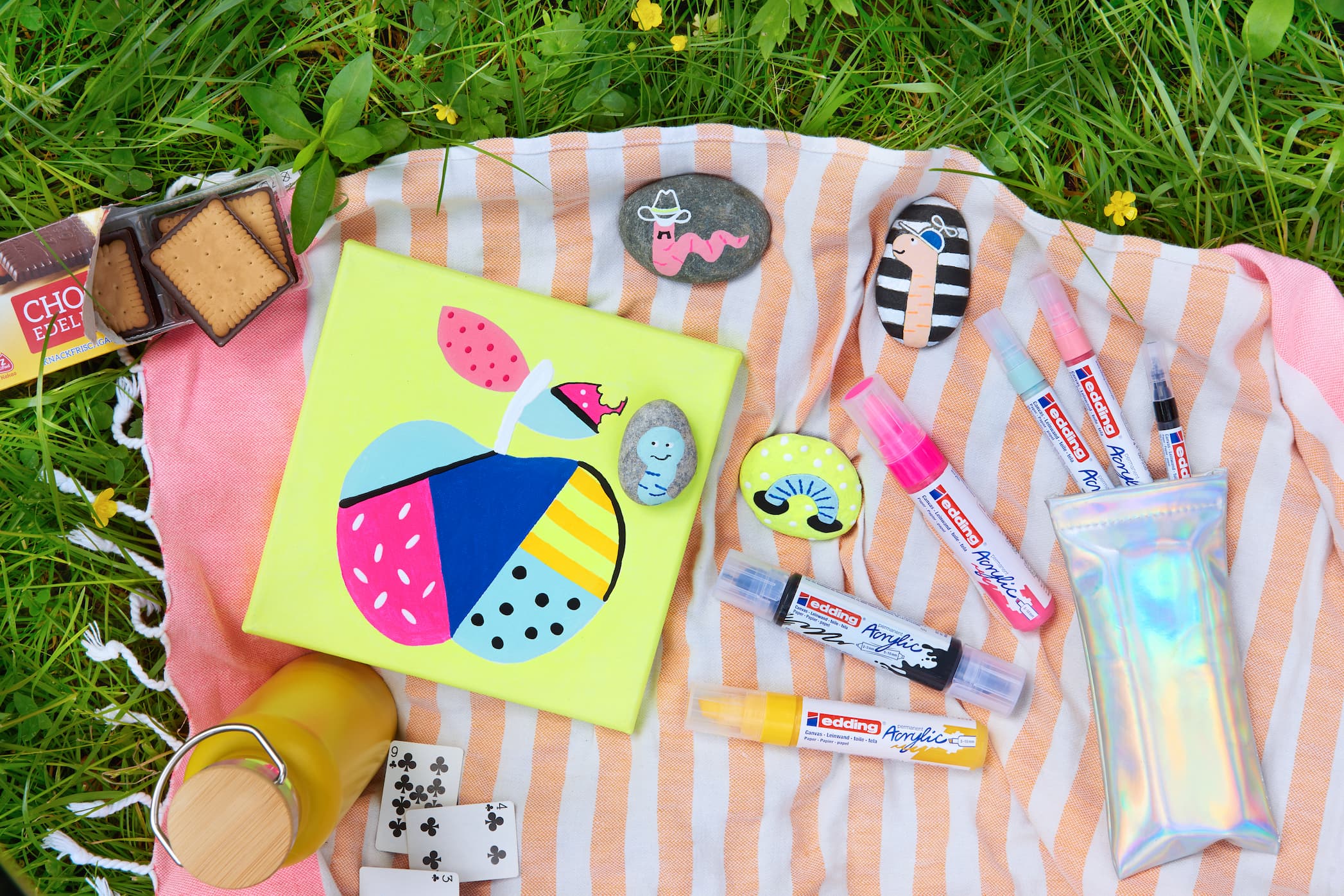 Eine lebendige Picknick-Szene mit einem farbenfrohen, abstrakten Design, erstellt mit edding 5000 Acrylmarker breit auf einem Notizbuch, verschiedenen skurrilen Abzeichen, aromatisierten Lippenbalsamen und einem Stück Schokolade, alles von edding.