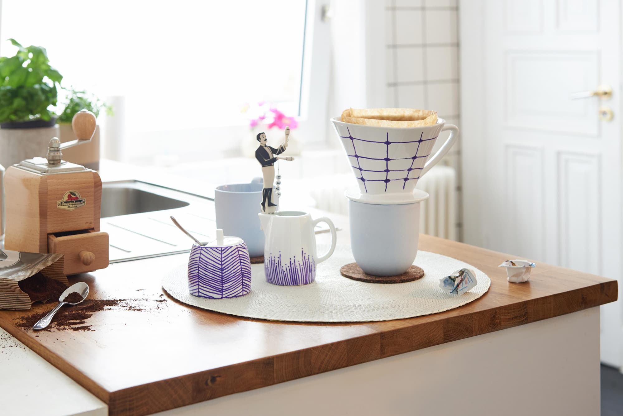 Eine Küchentheke mit Tassen, einer Kaffeemühle, einem Filter, einem Löffel, verschüttetem Kaffeesatz und Minifiguren – ideal für alle, die gerne Keramik mit dem edding 4200 Porzellanpinselstift von edding dekorieren und spülmaschinenfeste Designs kreieren.
