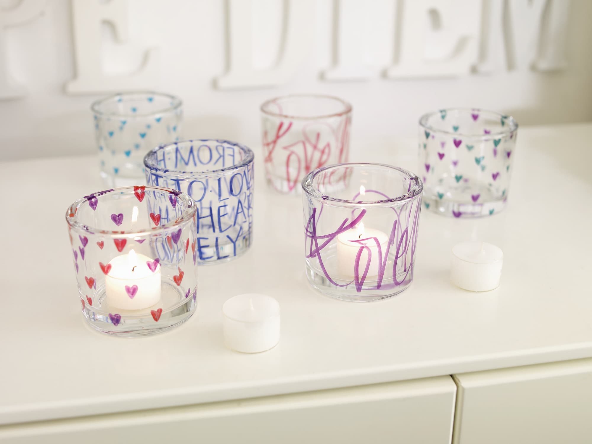 Sechs Kerzenhalter aus Glas, die mit dem edding 4200 Porzellanpinselstift von edding mit bunten Herzen, Wörtern und filigranen Motiven verziert sind, halten Teelichter auf einer weißen Fläche. Zwei unbeleuchtete Teelichter stehen davor; im Hintergrund ist ein Wandbild zu sehen.