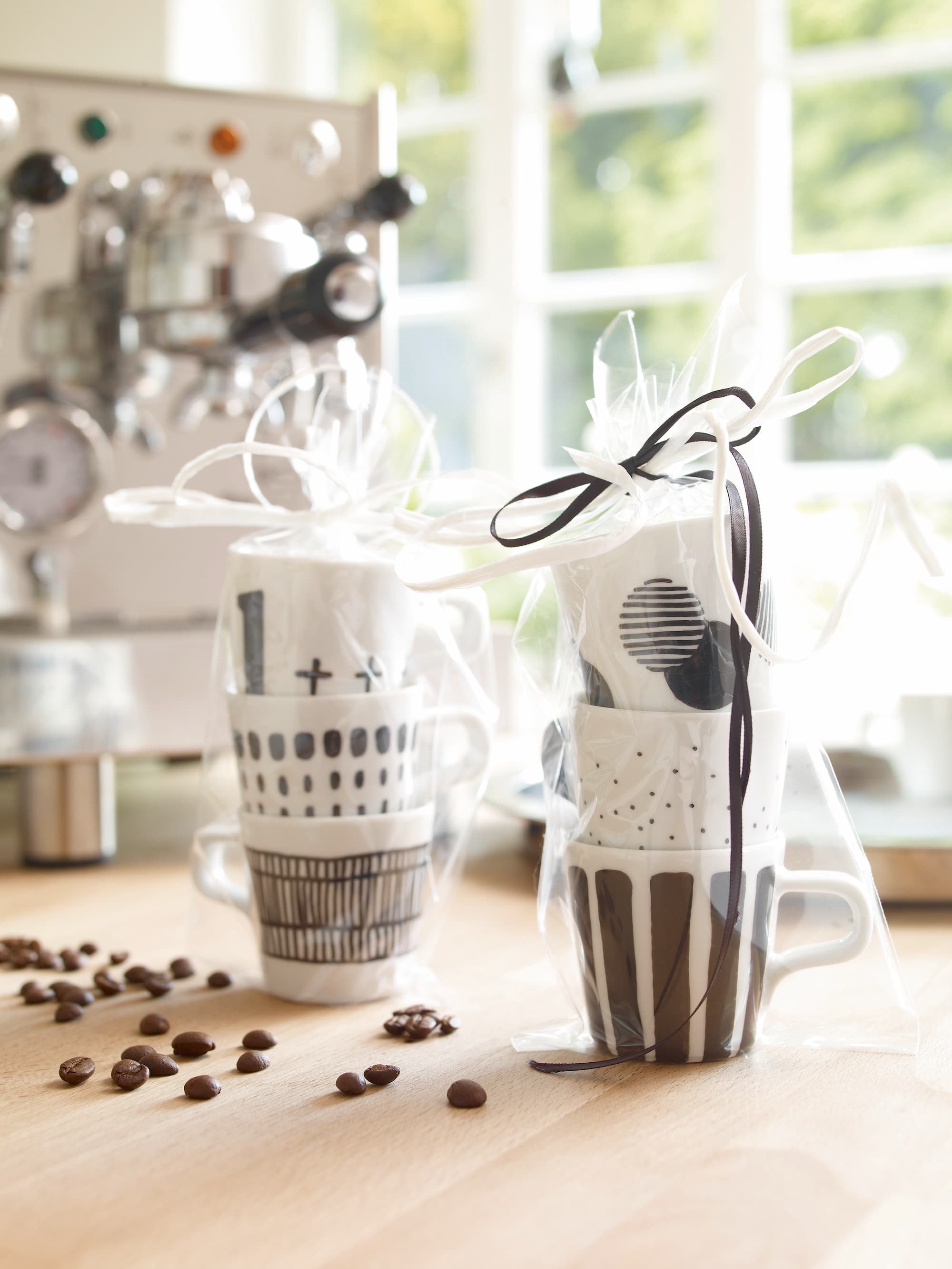 Zwei Kaffeetassen mit schwarz-weißen geometrischen Mustern – perfekt zum Keramikdekorieren mit dem edding 4200 Porzellanpinselstift von edding und spülmaschinenfest – sind in Geschenktüten verpackt und stehen auf einer Holztheke mit Kaffeebohnen, einer Maschine und einem sonnenbeschienenen Fenster im Hintergrund.