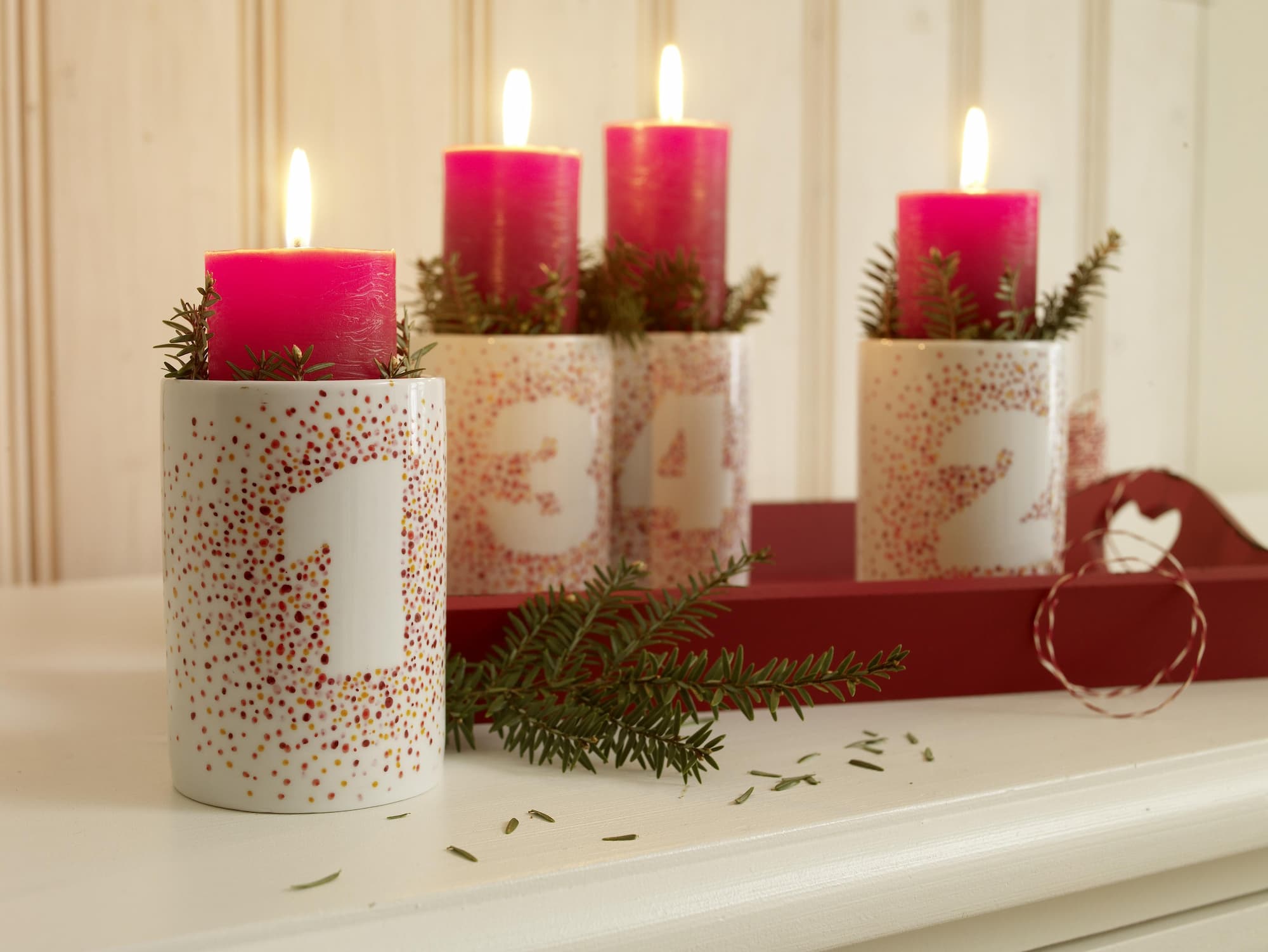 Vier rosa Kerzen in weißen, spülmaschinenfesten Haltern, nummeriert von 1–4 und mit Grün dekoriert, stehen zur Adventszeit auf einem roten Tablett. Die Zahlen sind mit dem edding 4200 Porzellanpinselstift von edding geschrieben.