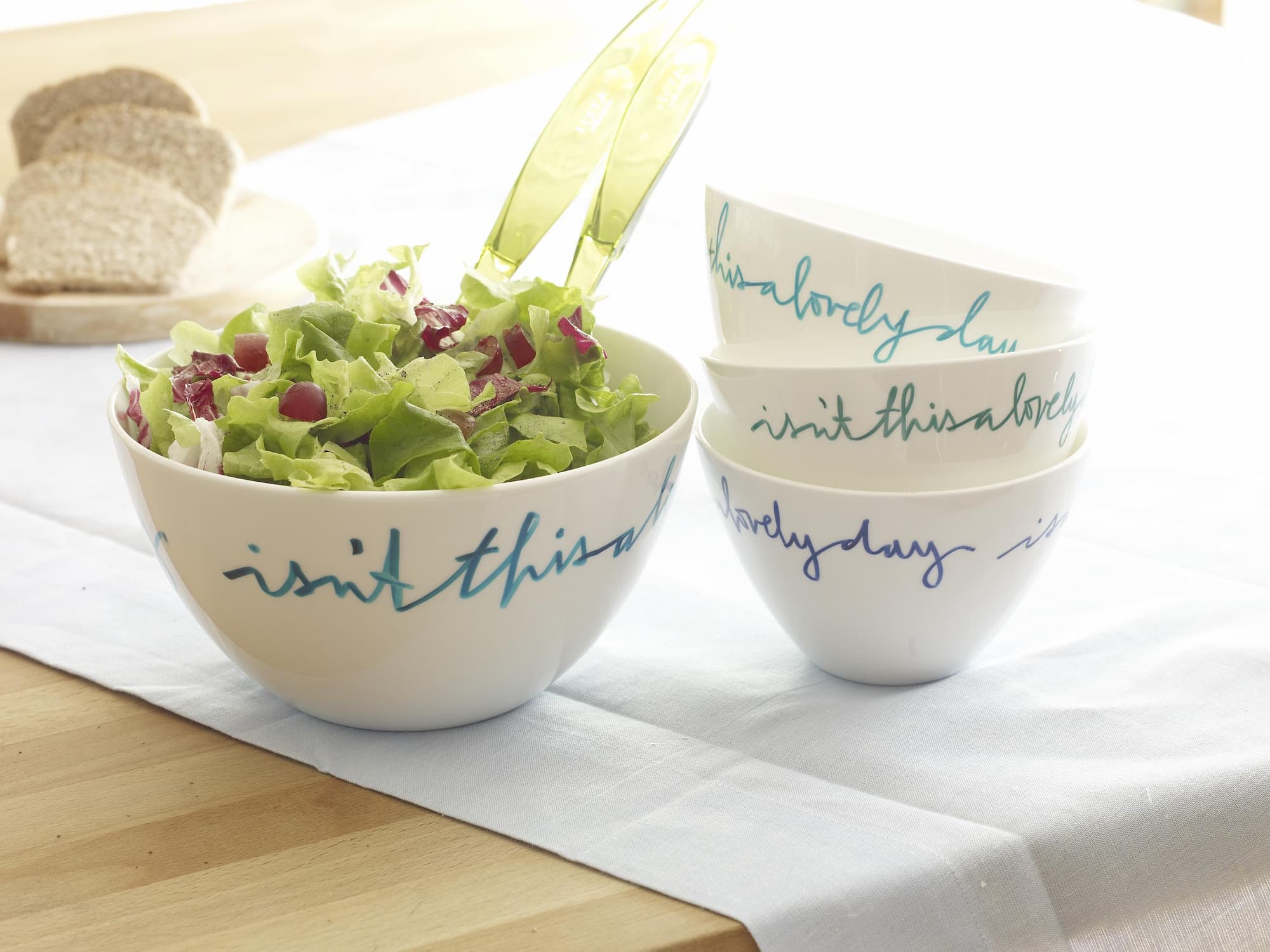 Eine Schüssel mit frischem Salat und einer grünen Salatzange steht neben gestapelten Schüsseln mit Schreibschrift, die wahrscheinlich mit dem edding 4200 Porzellanpinselstift von edding verziert wurden. Das Geschirr ist spülmaschinenfest, im Hintergrund sind Brotscheiben zu sehen.