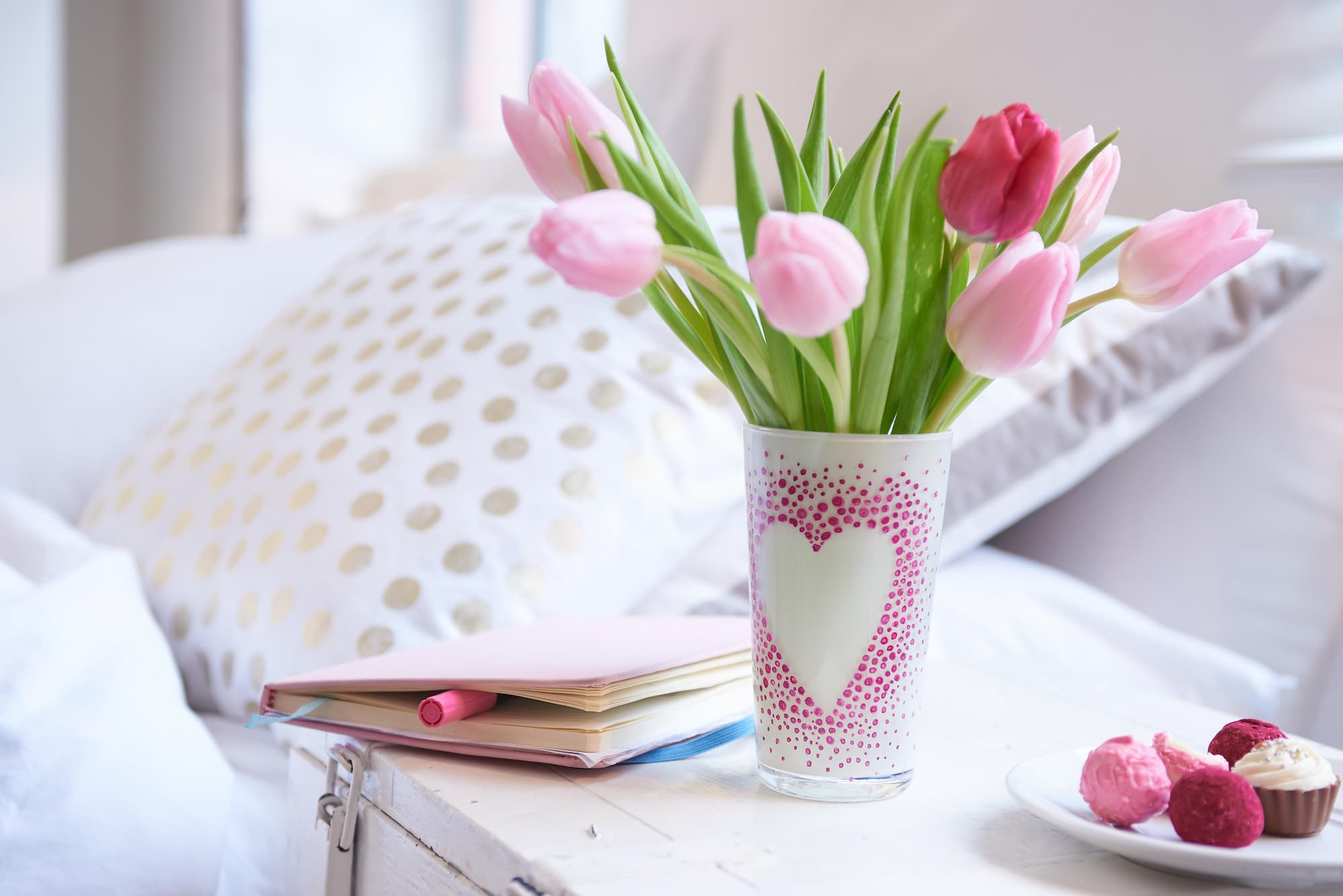 Rosa und rote Tulpen in einer weißen Vase mit Herz, dekoriert mit dem edding 4200 Porzellanpinselstift von edding für spülmaschinenfeste Details, auf einem Tisch neben einem gepunkteten Kissen, einem Notizbuch mit rosa Stift und rosa-weißen Süßigkeiten.