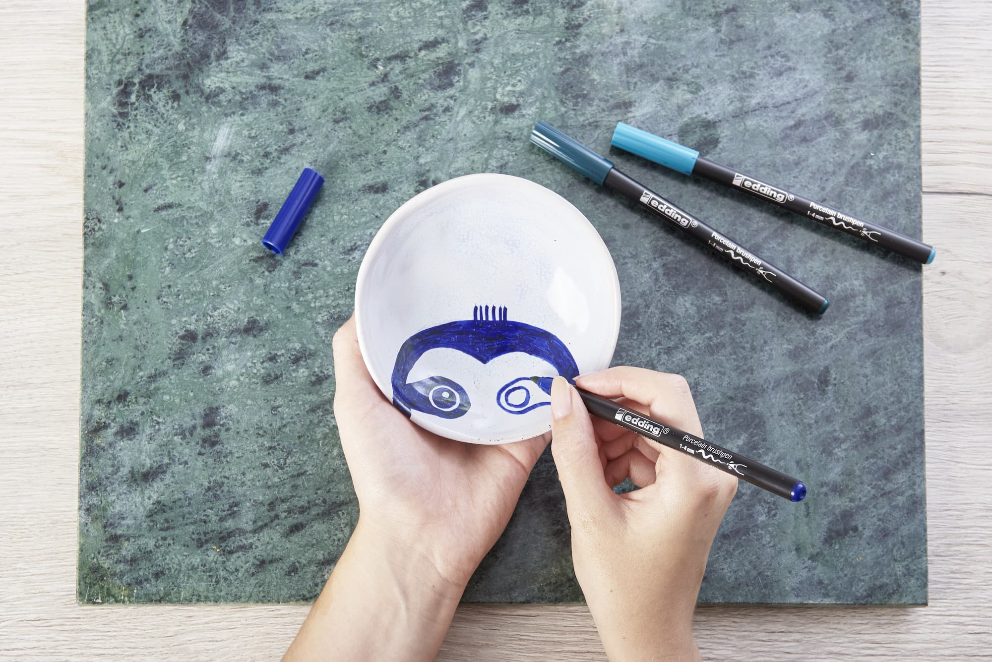 Mit dem edding 4200 Porzellanpinselstift von edding verziert jemand eine weiße Keramikschale mit blauen und schwarzen Cartoon-Gesichtern, während zwei weitere edding-Marker und eine Kappe auf der grünen strukturierten Oberfläche daneben liegen.
