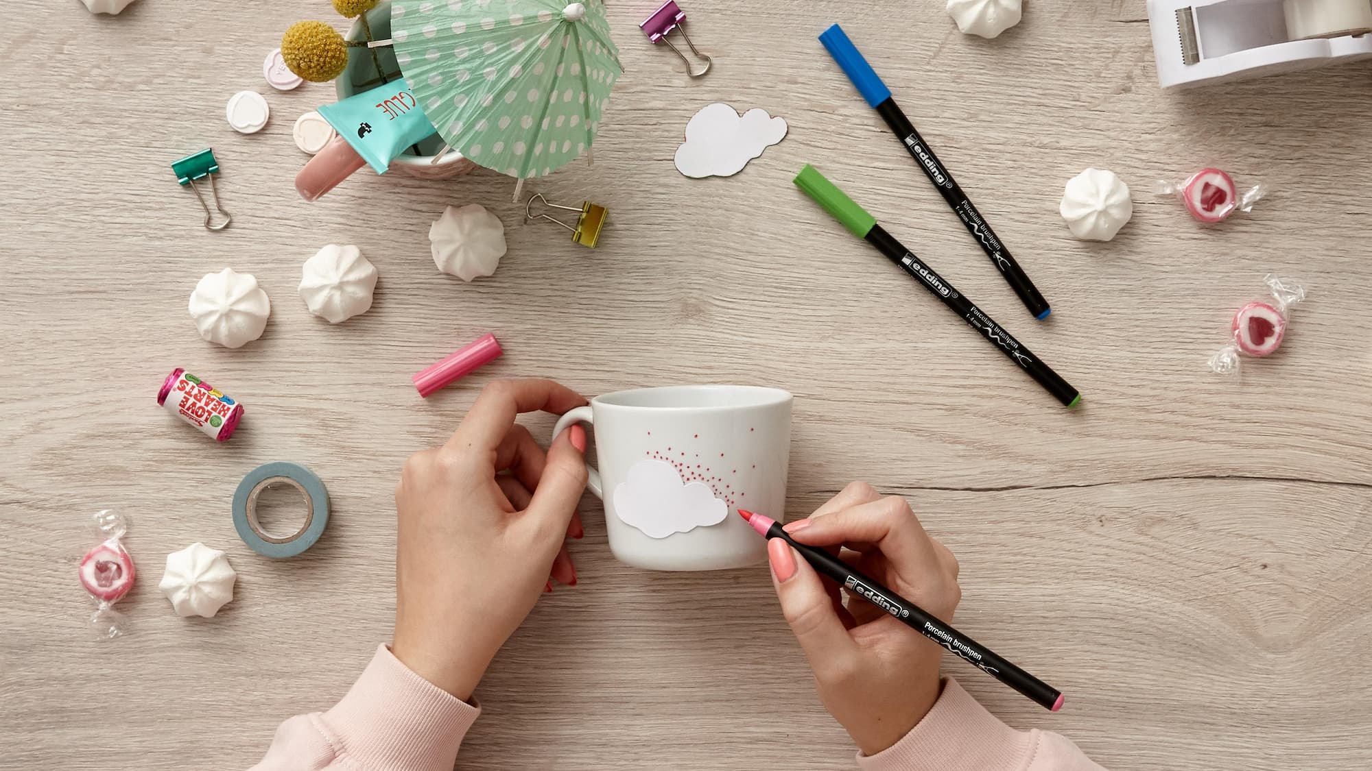 Jemand verziert eine weiße Tasse mit einem Wolkenmotiv und verwendet dazu den rosa edding 4200 Porzellanpinselstift von edding. Bunte Stifte, Süßigkeiten, Büroklammern, Baisers, Klebeband und ein Cupcake-Topper-Schirm liegen beim Keramikdekorieren verstreut auf dem Holztisch.