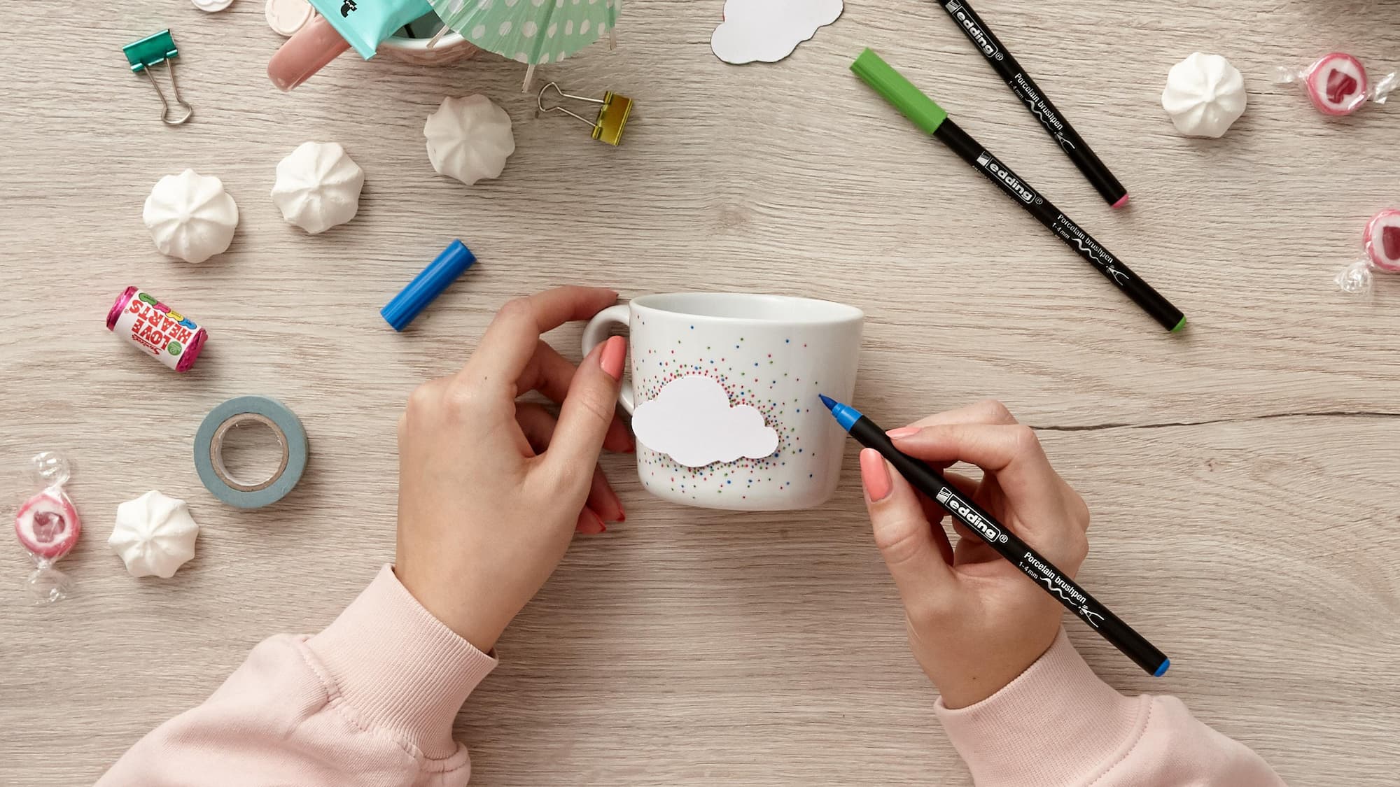 Hände halten eine weiße Tasse mit einem Wolkenaufkleber und bunten Punkten, umgeben von Markern, Klebeband, Süßigkeiten und Künstlerbedarf. Eine Hand verziert die Keramiktasse mit einem blauen edding 4200 Porzellanpinselstift von edding.