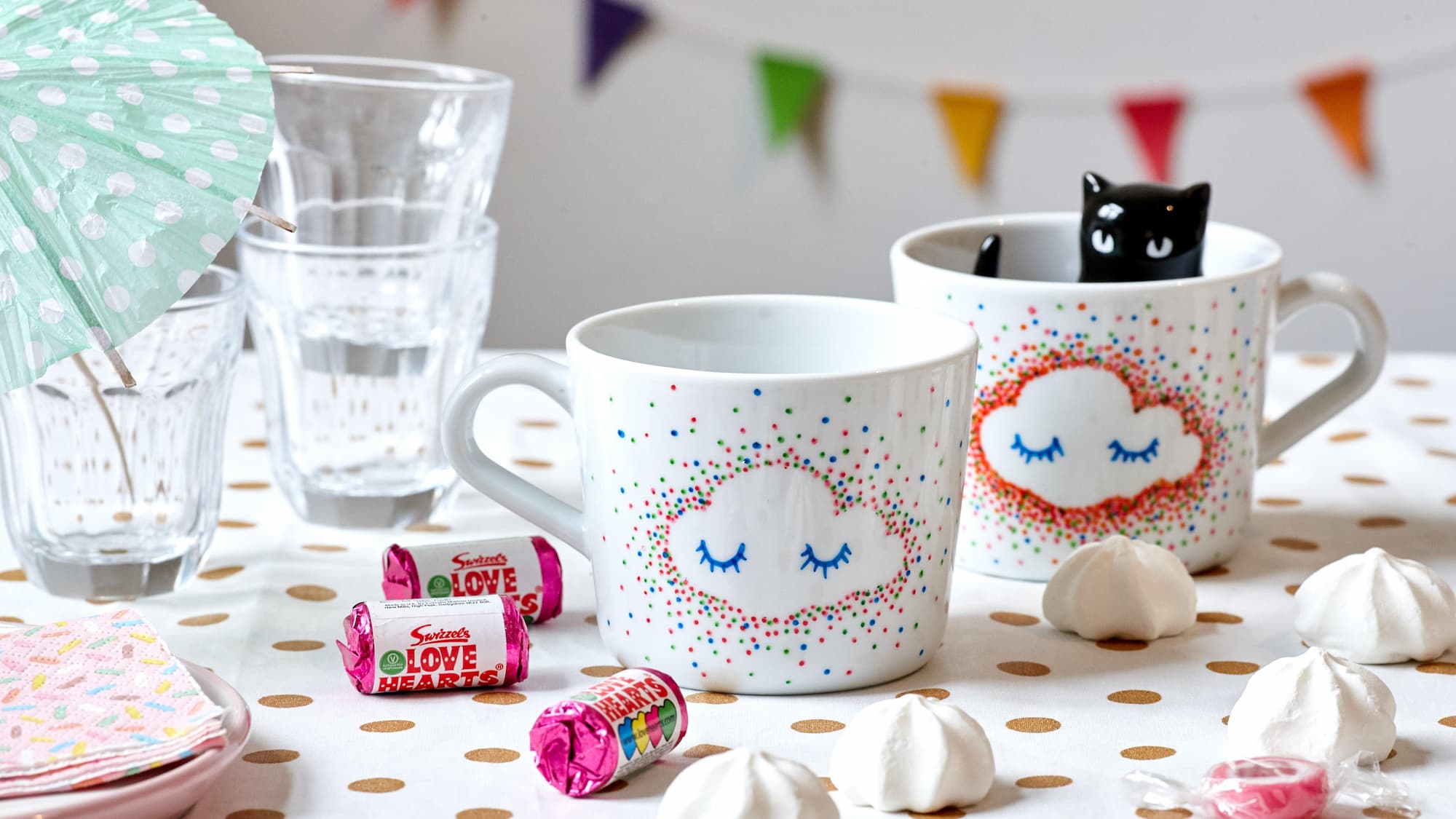 Zwei weiße Tassen, verziert mit bunten Wolkenmotiven, die mit dem edding 4200 Porzellanpinselstift von edding gestaltet wurden, stehen auf einer gepunkteten Tischdecke. In einer Tasse befindet sich eine schwarze Katzenfigur. Daneben stehen Gläser, Baisers, Süßigkeiten, Servietten und Wimpel.