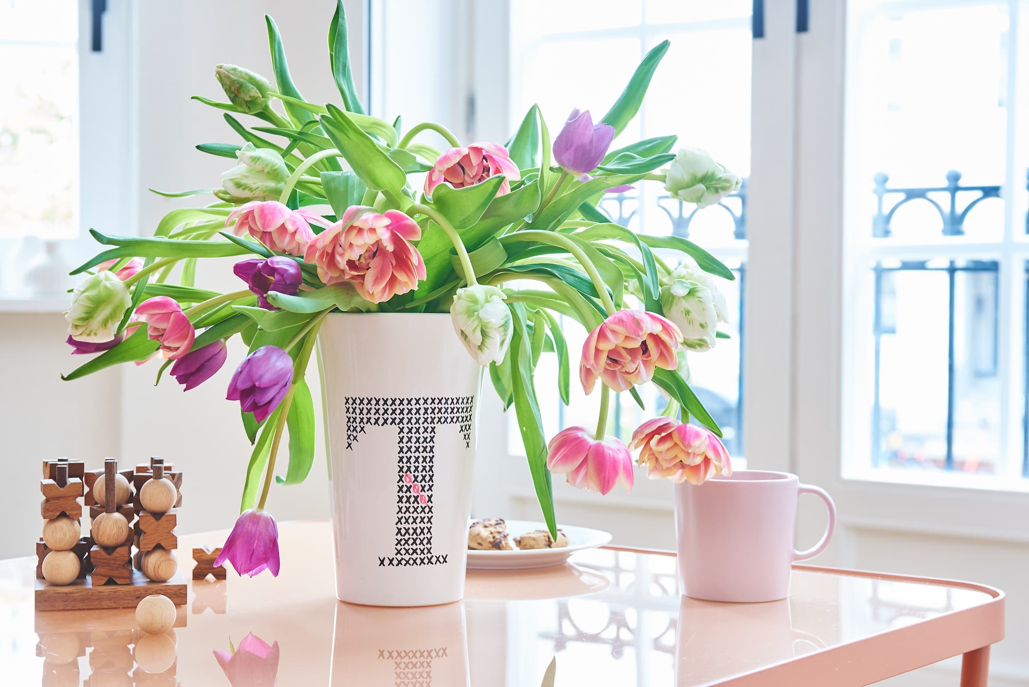 Ein edding 4200 Porzellanpinselstift von edding steht auf einem Tisch mit einer weißen Vase mit Tulpen, einer rosa spülmaschinenfesten Tasse, Keksen auf einem Teller und einem Holzpuzzle, während Sonnenlicht durch die hellen Fenster dahinter strömt.