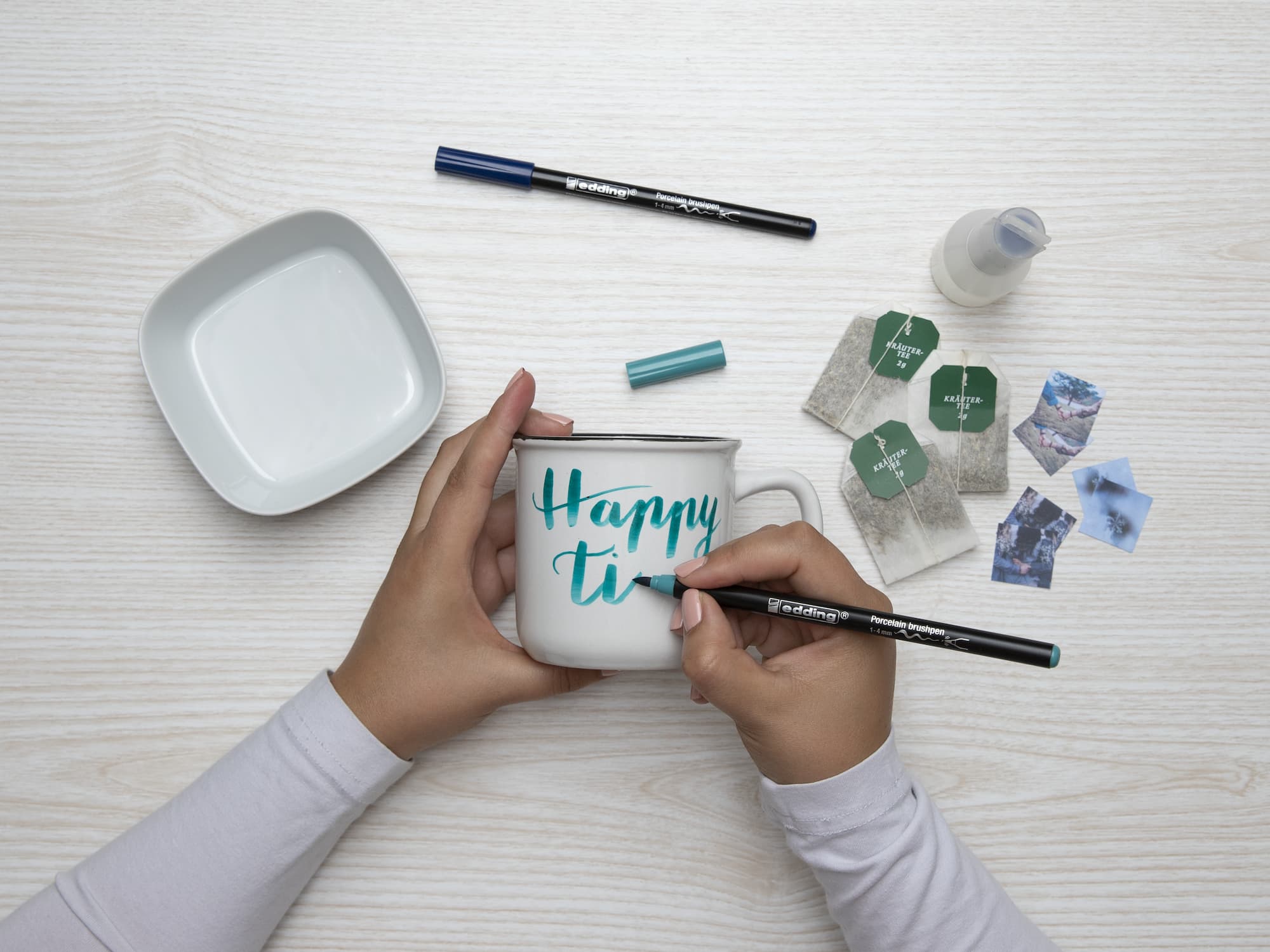 Jemand verziert eine weiße Tasse mit dem blauen Motiv „Happy Times“ mit dem edding 4200 Porzellanpinselstift von edding. Auf einer hellen Holzoberfläche befinden sich ein weißes Geschirr, Marker, Teebeutel, Fotos und eine Flasche. Das Motiv ist spülmaschinenfest und sorgt für langanhaltende Freude.