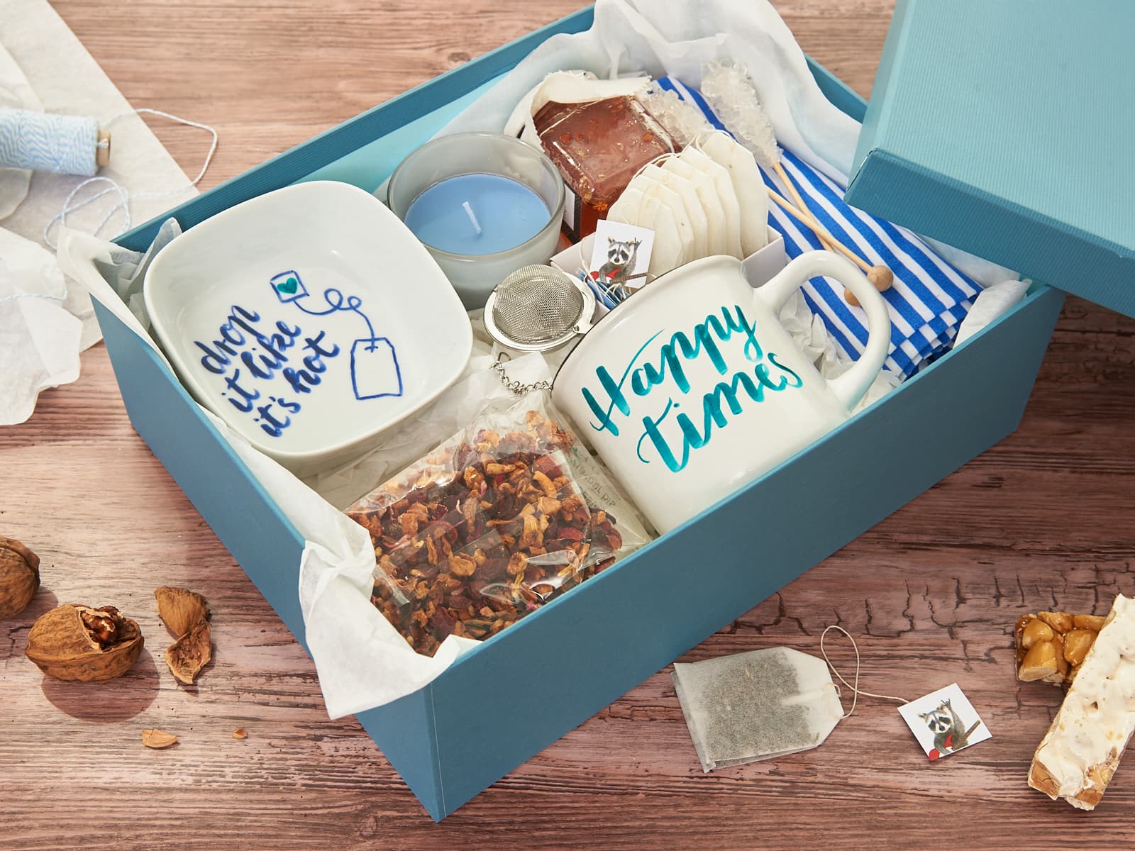 Eine blaue Geschenkbox mit einem edding 4200 Porzellanpinselstift von edding, einer Tasse mit der Aufschrift „Don’t tick it’s hot“, einer Kerze, Teebeuteln, Trockenfrüchten, Seife und einem gestreiften Beutel – arrangiert auf Seidenpapier auf einer Holzoberfläche.