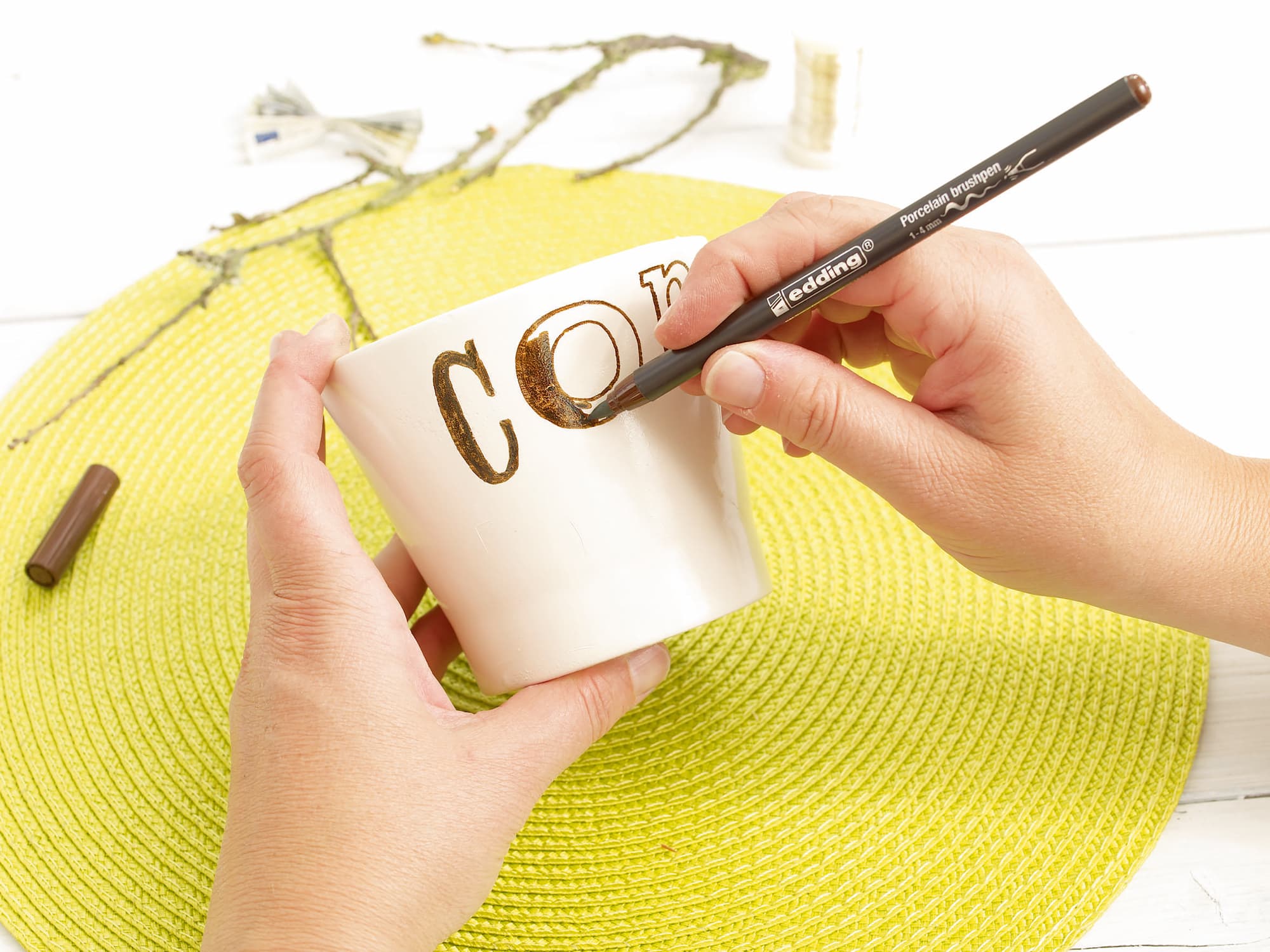 Mit einem edding 4200 Porzellanpinselstift verziert jemand eine weiße Keramiktasse mit dem Schriftzug „CON“ und hält sie über ein gelbes Tischset auf einem weißen Tisch. Nach dem Trocknen und Einbrennen ist das Motiv spülmaschinenfest.