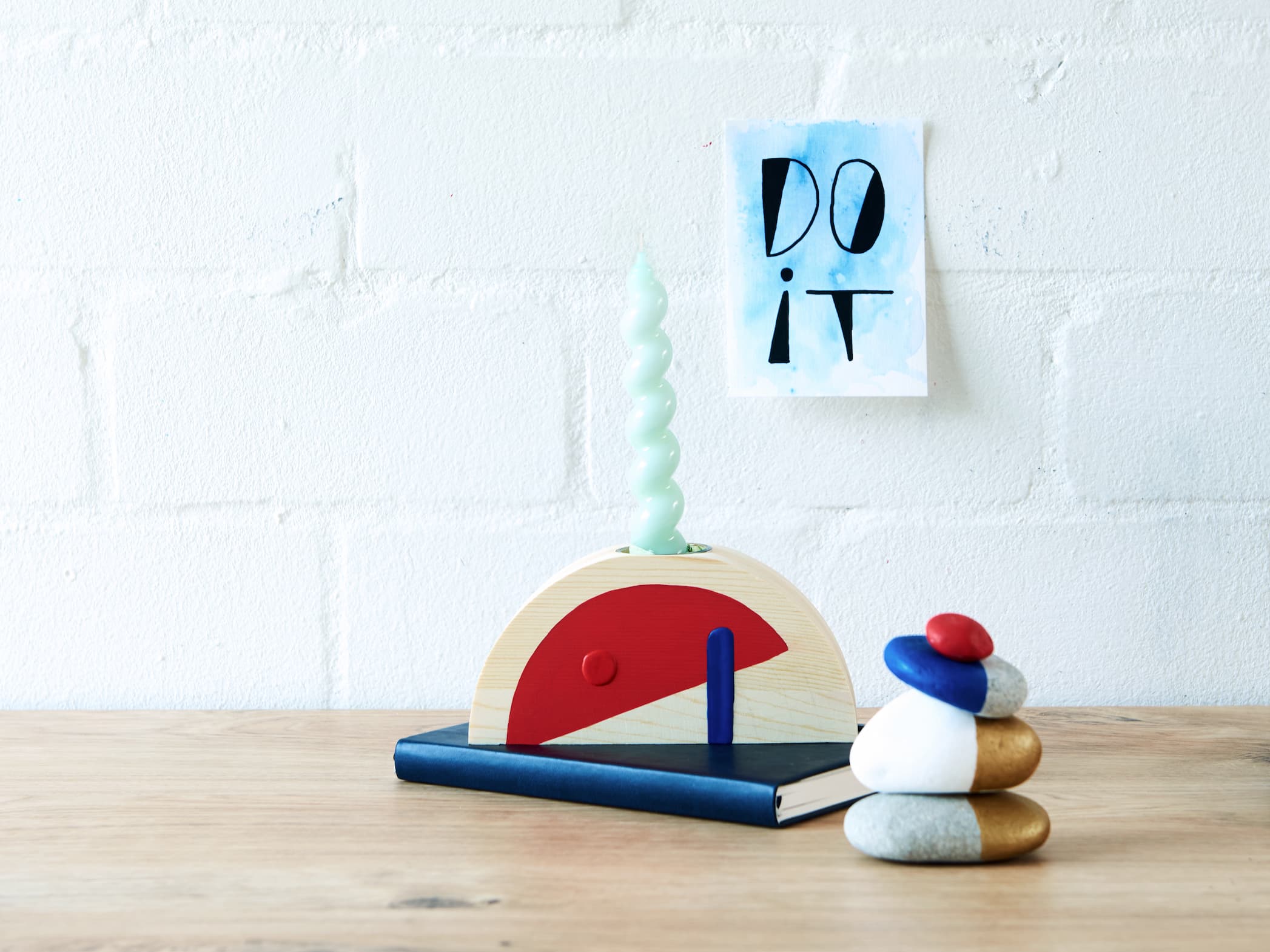Ein minimalistischer Schreibtischaufbau mit einem abstrakten hölzernen Schreibtischspielzeug, einer Spiralkerze, einem Stapel balancierender Steine und einem Motivationsposter mit den Worten „do it“ in edding 5000 Acrylmarker breit.