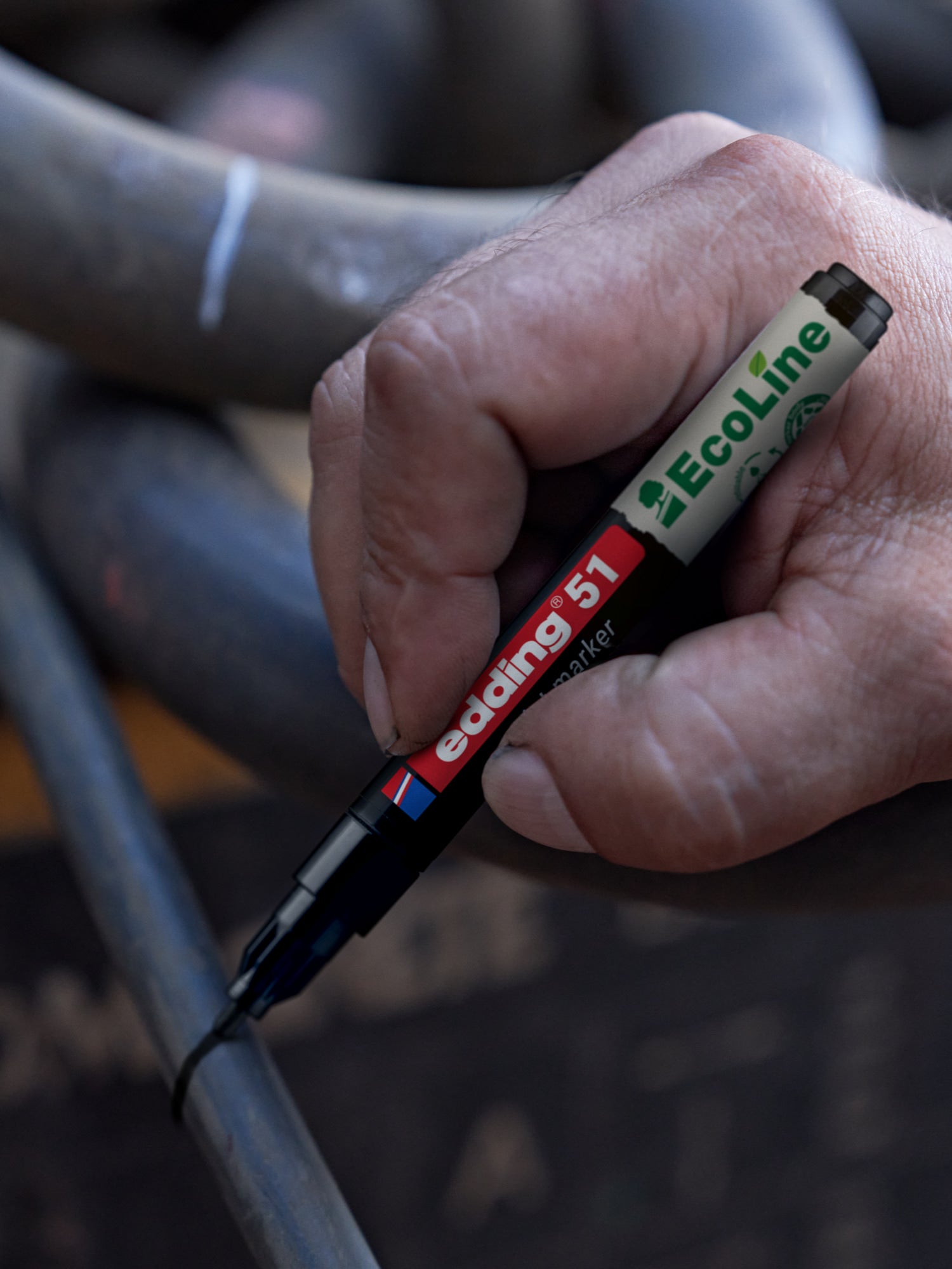 Eine Hand zeichnet mit einem schwarzen Edding 51 EcoLine Marker eine Linie auf ein Metallrohr, mit verschlungenen Rohren im Hintergrund.