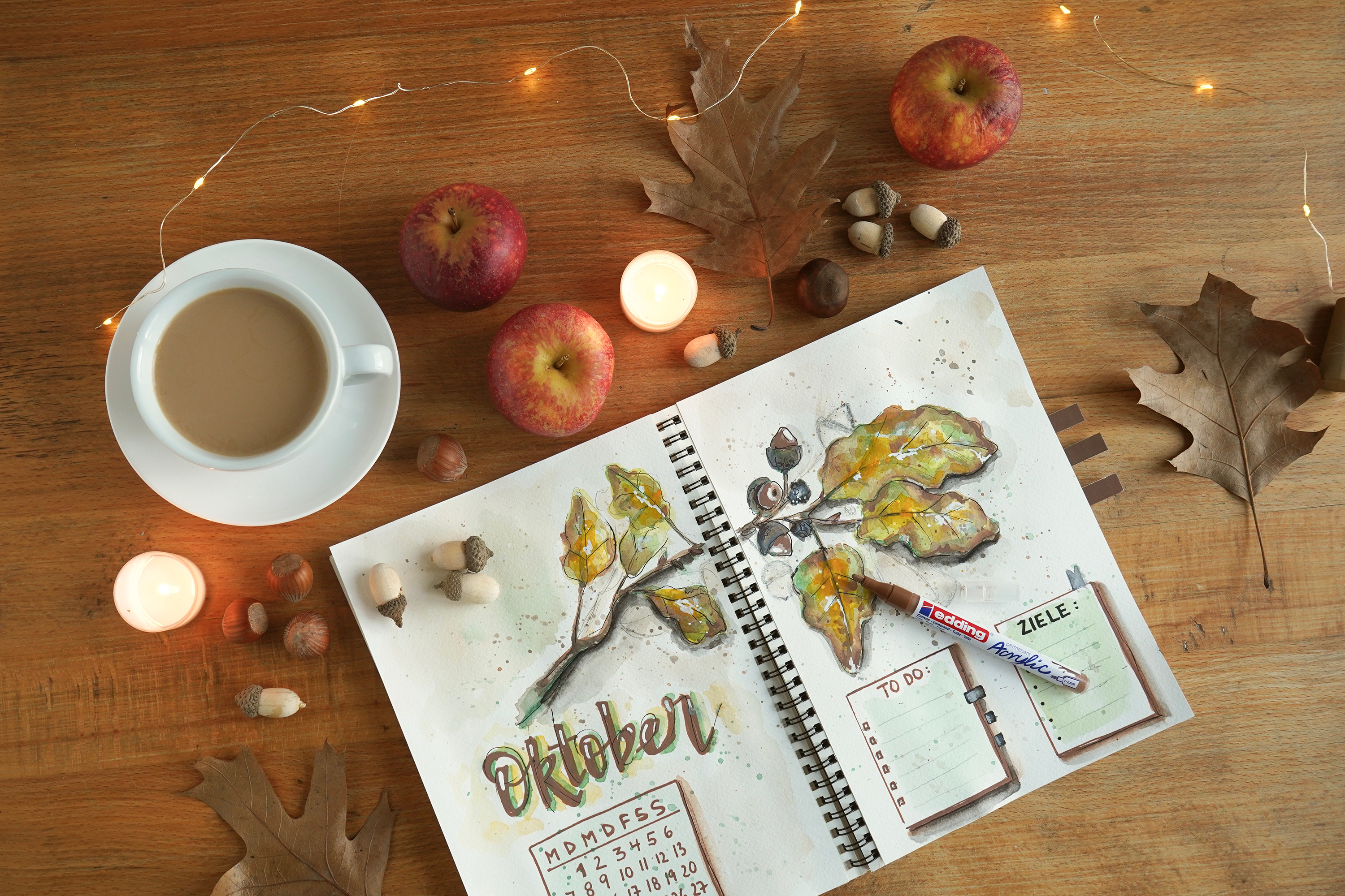 Eine gemütliche Herbstszene mit einem aufgeschlagenen Kunstjournal für ein edding 5300 Acrylmarker fein 5er-Set-Gemälde zum Thema Oktober, umgeben von kleinen Kerzen, frisch gepflückten Äpfeln, Eicheln und trockenen Blättern auf einem Holztisch.