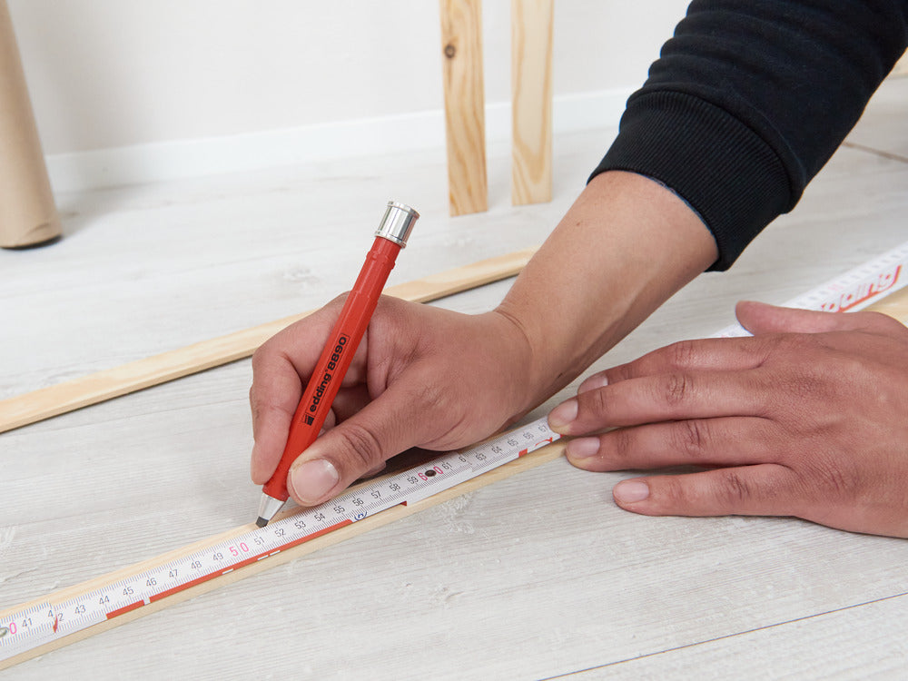 Mit dem edding Heimwerker-Bundle von edding SHOP wird mit einem roten Zimmermannsbleistift und Maßband auf einer hellen Holzfläche ein Maß auf einem Holzstab markiert.