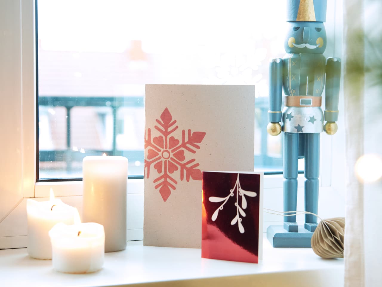 Eine gemütliche Weihnachtsdekoration mit einer Nussknackerfigur, die Wache neben Grußkarten steht, Kerzen, die einen warmen Schein verbreiten, und das alles vor einem sanft beleuchteten Fenster mit edding 1340 metallischen Pinselstift-Dekorationen.