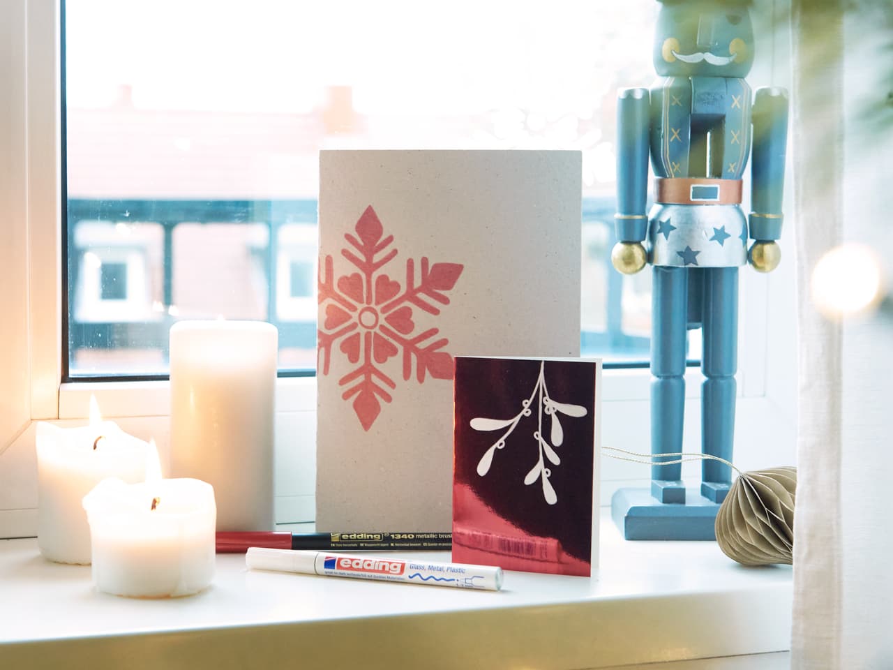 Ein gemütliches Festtags-Setup mit brennenden Kerzen, einer handgefertigten Grußkarte mit einem Schneeflocken-Design, erstellt mit hochwertiger wasserbasierter Tinte und edding 1340 metallic Pinselstift, Bastelmaterialien.
