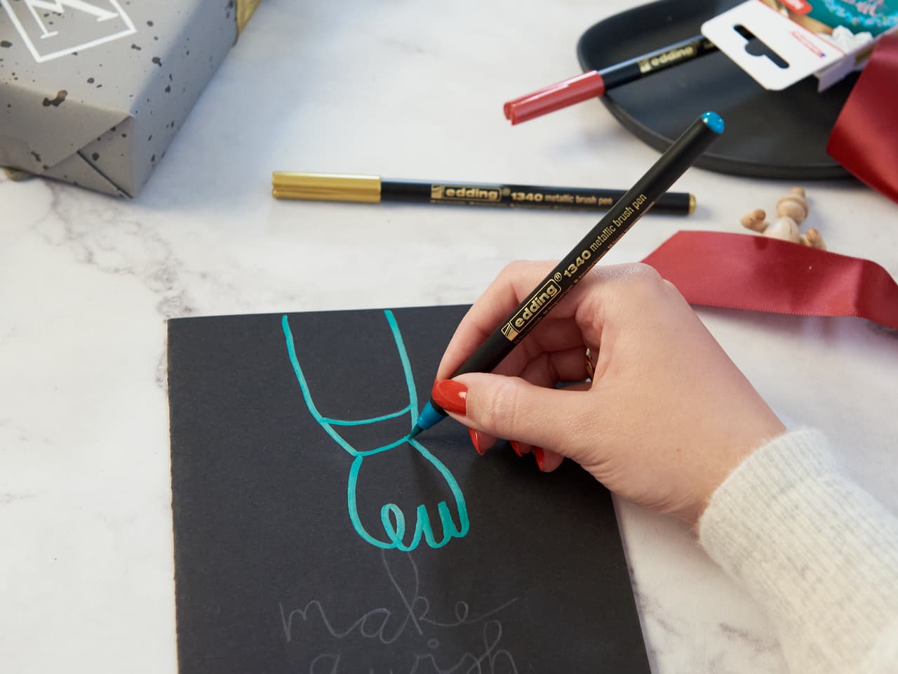 Eine Hand hält einen edding 1340 metallic Pinselstift mit wasserbasierter Tinte und zeichnet eine elegante türkisfarbene Schleife auf ein schwarzes Blatt Papier, während künstlerisches Zubehör verstreut ist.