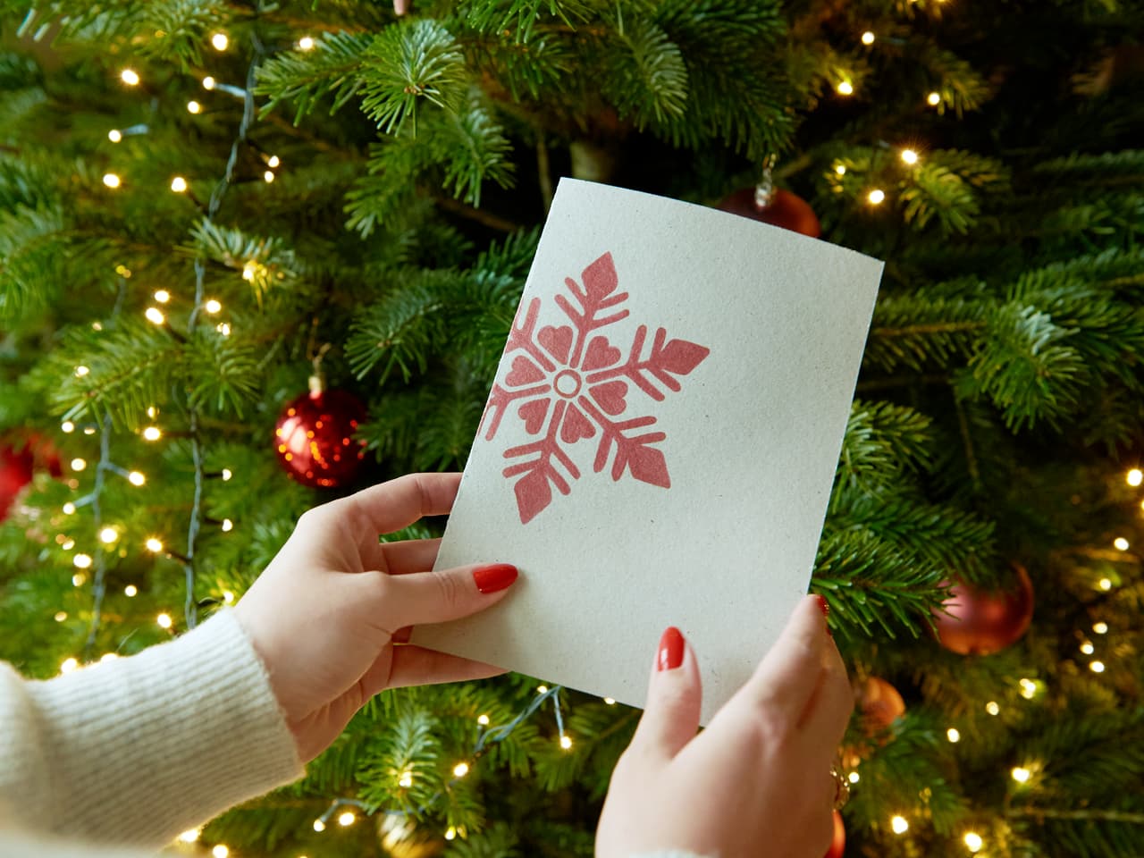 Hände präsentieren eine festliche Weihnachtskarte mit Schneeflocken-Design, erstellt mit einem edding 1340 Metallic-Pinselstift, vor dem Hintergrund eines geschmückten Weihnachtsbaums mit funkelnden Lichtern.
