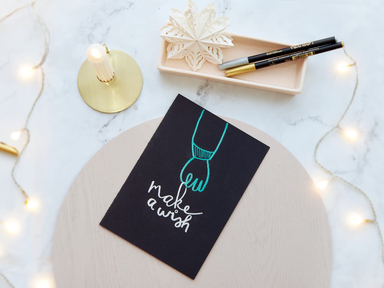 Ein charmantes Flatlay mit einer „Wünsch dir was“-Grußkarte, begleitet von einem edding 1340 Metallic Pinselstift mit wasserbasierter Tinte, einer zarten Schneeflockendekoration und funkelnden Lichterketten.