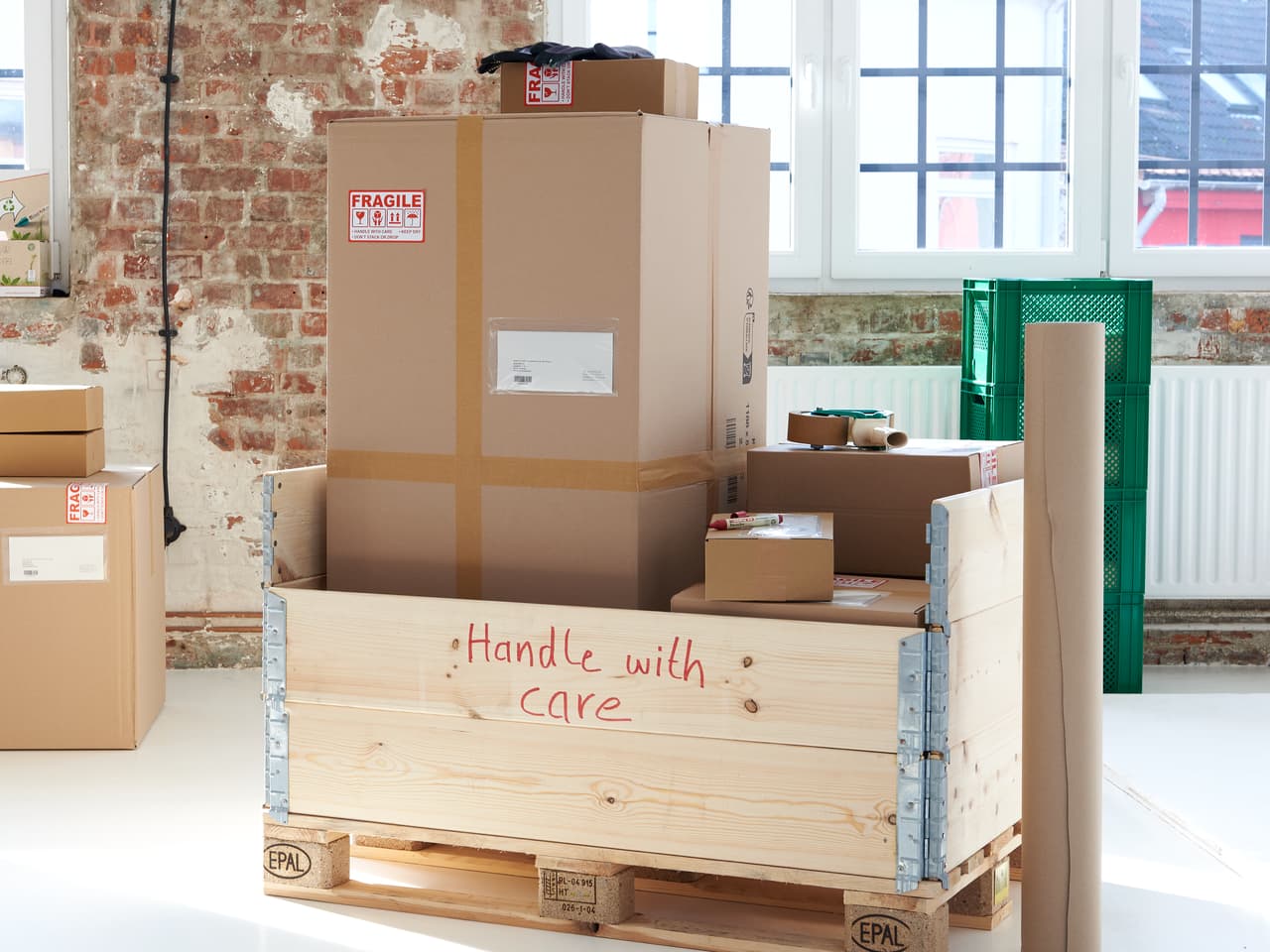 Ein geräumiger Raum mit großen Fenstern zeigt eine Sammlung verpackter Kartons, von denen einige zerbrechlich sind und ordentlich auf einer Holzpalette platziert sind und auf den Versand warten. Auf der einen Seite befindet sich ein umweltfreundliches edding 22 EcoLine Permanentmarker 4er-Set, ordentlich beschriftet mit dem Markennamen „edding“.