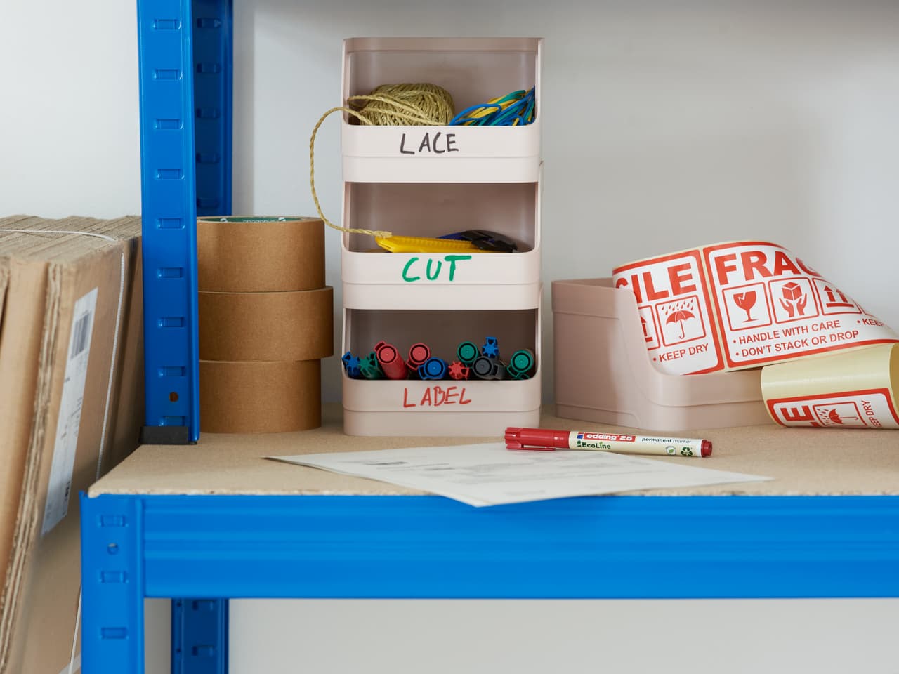 Eine organisierte Versandstation mit beschrifteten Behältern, edding 25 EcoLine Permanentmarkern und Versandetiketten auf einem Regal.