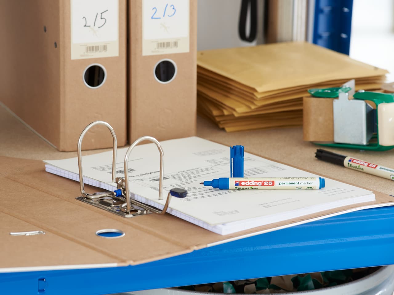 Ein offener Ringordner mit Dokumenten auf einem Schreibtisch, umgeben von BĂĽromaterialien, darunter edding 25 EcoLine Permanentmarker, Stiften und Aufbewahrungsboxen aus Pappe.