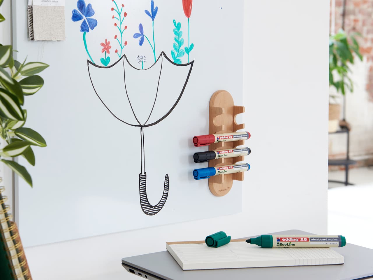 Ein kreativer Arbeitsplatz mit einer skurrilen Wandzeichnung einer Topfpflanze, mit farbenfrohen edding 28 EcoLine Whiteboardmarkern, die magnetisch an einem Halter aus recyceltem Material befestigt sind, und einem Notizblock mit einem.