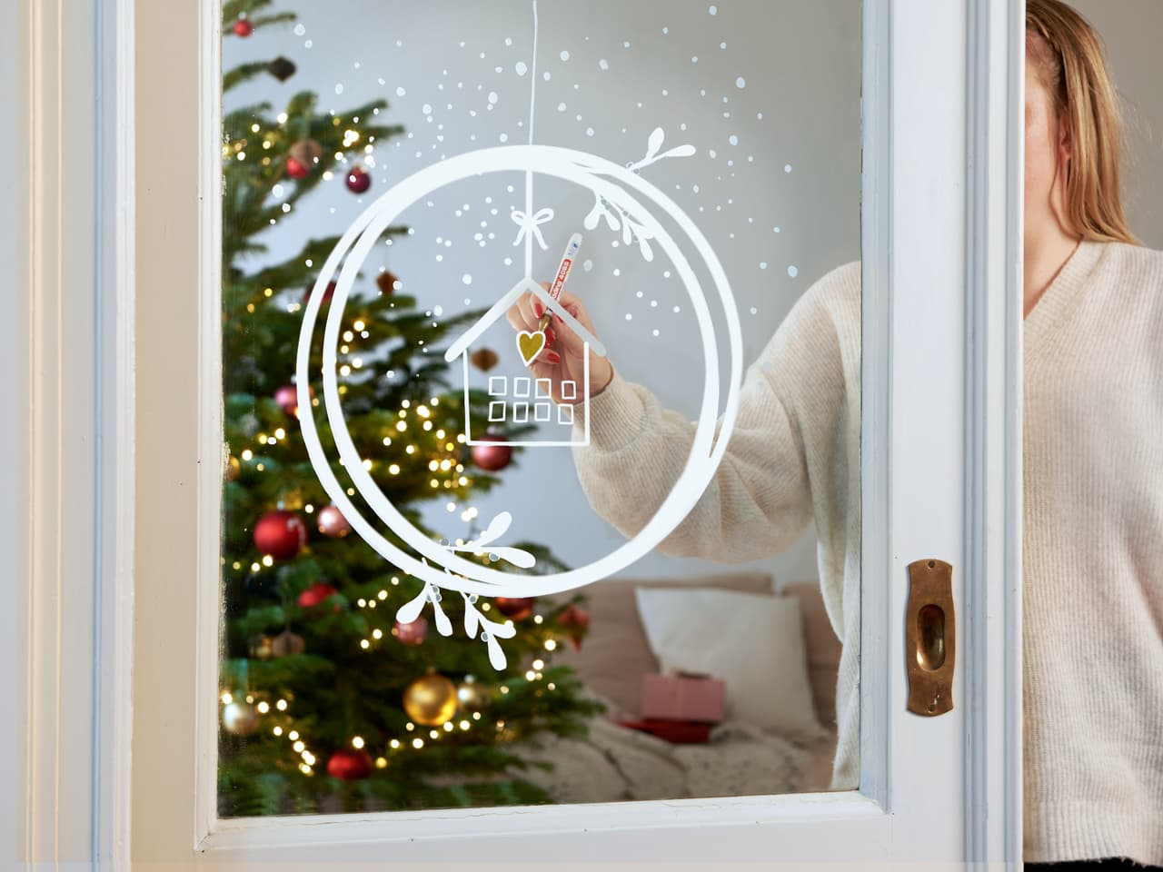Eine Person hält mit einem edding 4085 Kreidemarker eine Hausdekoration aus Papier mit einem präzisen Design an das Fenster, im Hintergrund verschwimmt ein festlicher Weihnachtsbaum, der mit Lichtern und Ornamenten geschmückt ist.