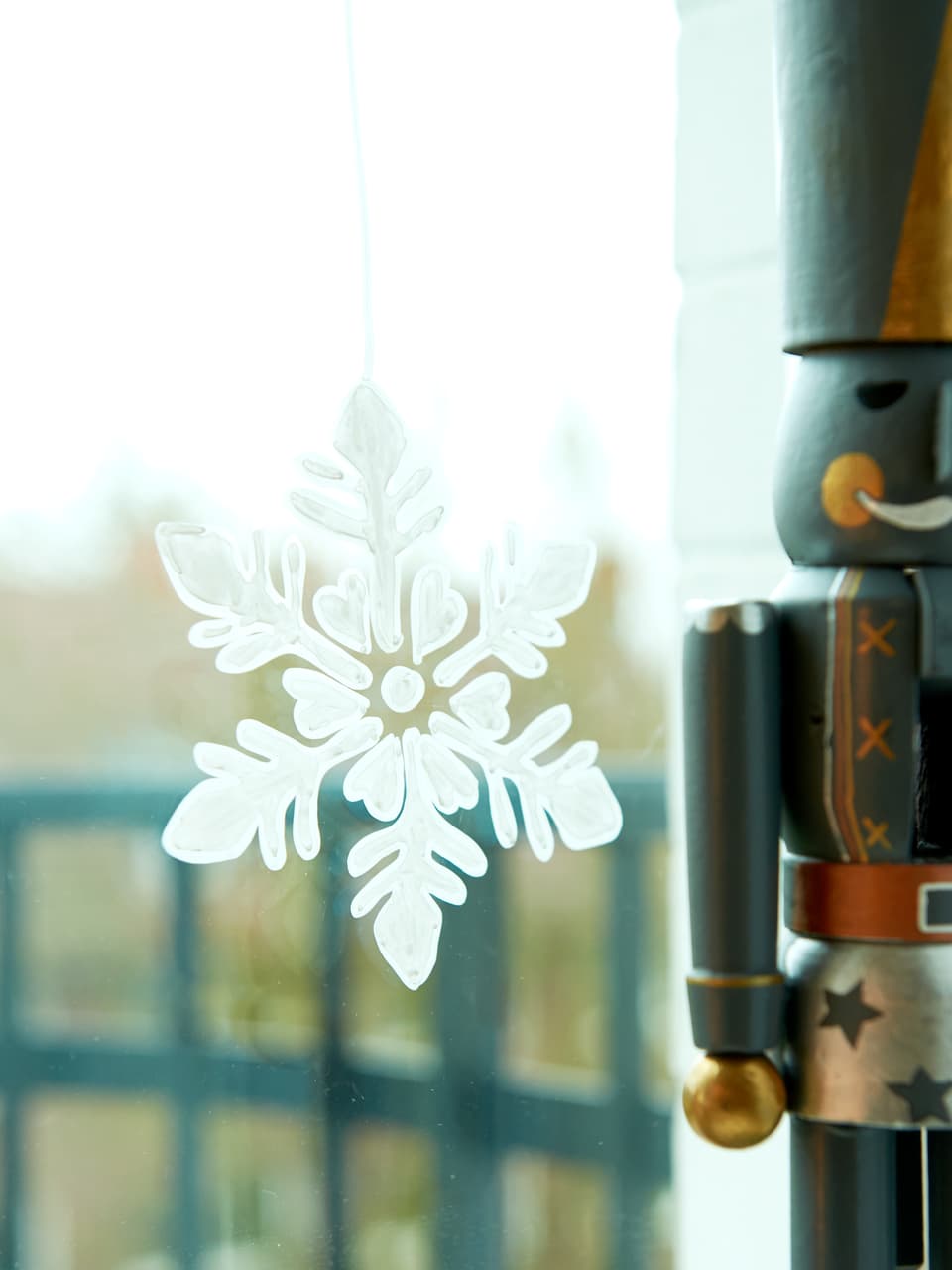 Ein zartes Schneeflockenornament, mit präziser Gestaltung mit dem edding 4085 Kreidemarker aus einem edding Kreidemarker erstellt, hängt neben einem verschwommenen Nussknacker an einem Fenster.