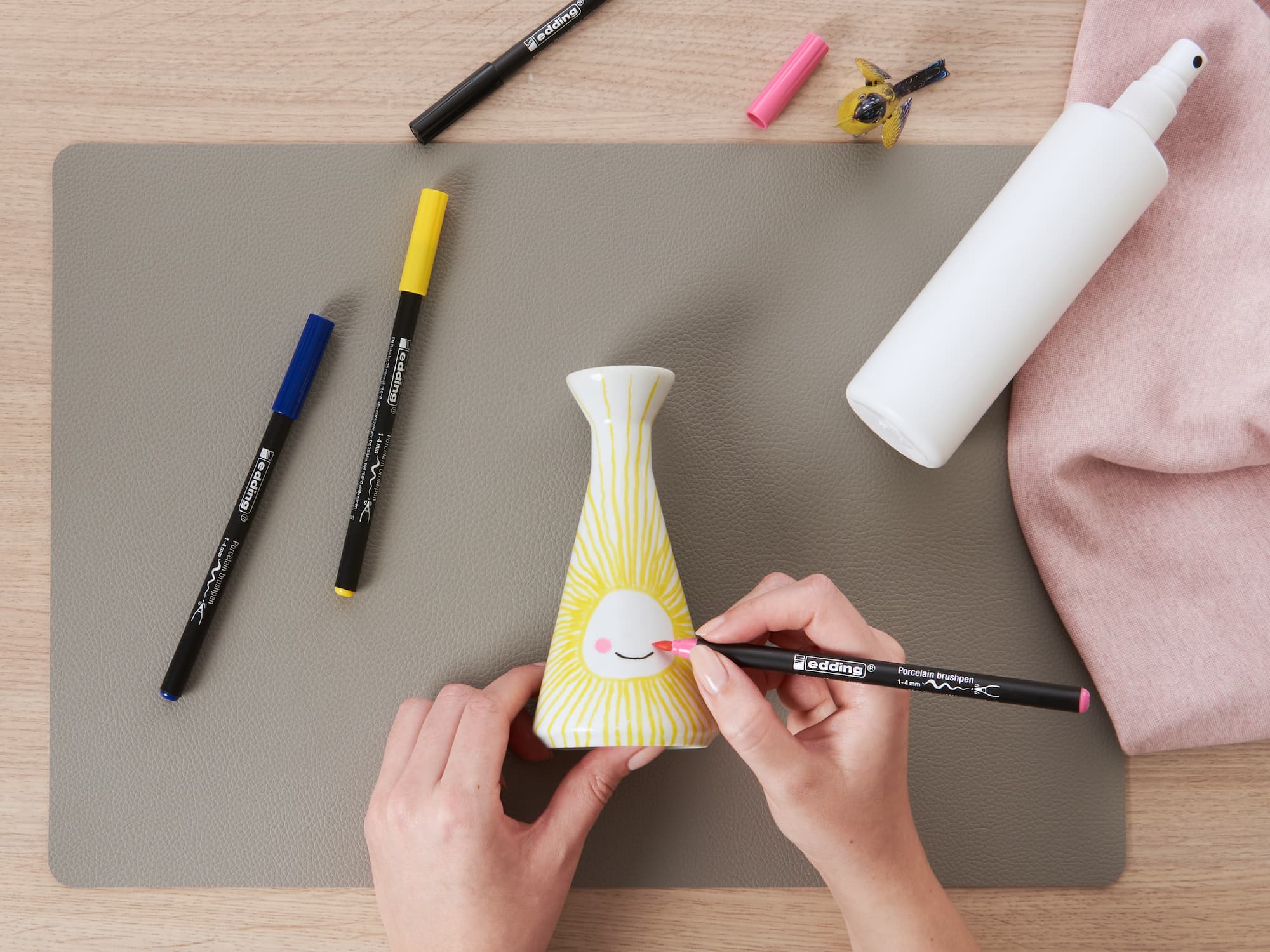 Jemand verziert eine weiße Vase mit gelben Linien und einer kleinen rosa Form mit dem edding 4200 Porzellanpinselstift von edding. Weitere Marker, eine Sprühflasche und ein rosa Tuch stehen auf dem Tisch – ideal für Keramik-Dekor-Fans.