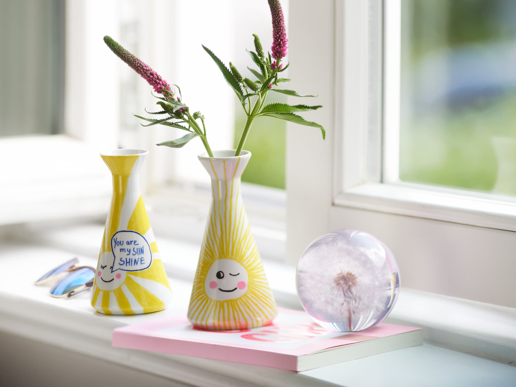 Zwei fröhliche Vasen mit Sonnenmotiven, dekoriert mit dem edding 4200 Porzellanpinselstift von edding, stehen auf einer Fensterbank – eine mit lila Blumen, die andere mit der Aufschrift „You are my sunshine“. Beide sind spülmaschinenfest. Ein Pusteblumen-Briefbeschwerer und eine Sonnenbrille runden die Szene ab.