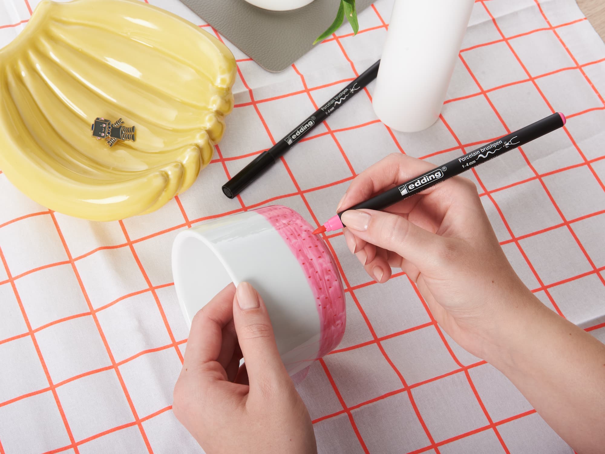 Mit einem edding 4200 Porzellanpinselstift von edding verziert jemand eine weiße Keramiktasse mit rosa Motiven auf einem roten Gittertuch; daneben stehen zwei schwarze Marker und eine gelbe bananenförmige Schale. Das Motiv ist spülmaschinenfest.