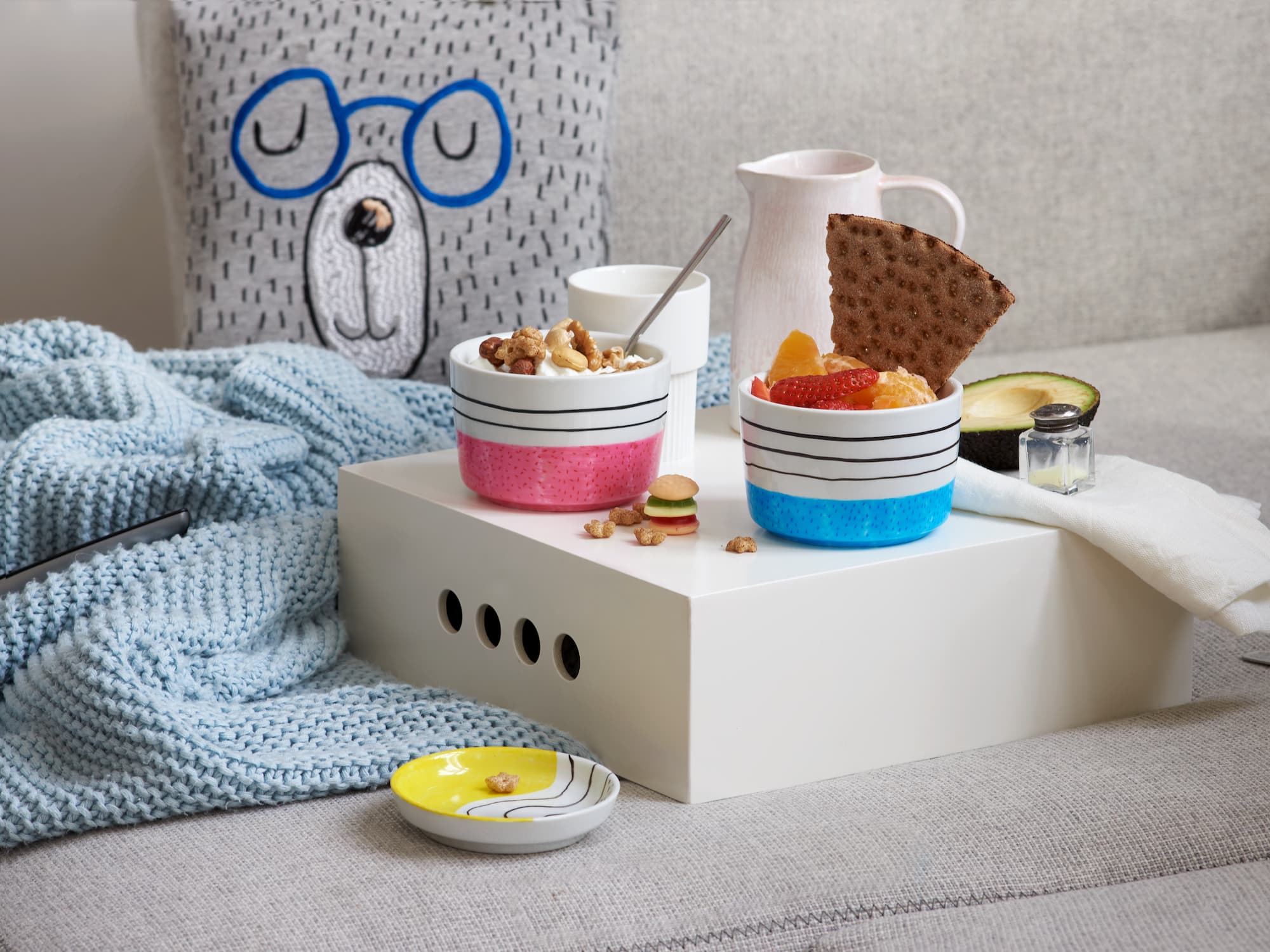 Zwei Snackschalen – eine mit Popcorn und Nüssen, die andere mit Obst und Knäckebrot – stehen auf einem weißen Tablett. Charmante Akzente mit dem edding 4200 Porzellanpinselstift von edding sorgen für Flair, während eine blaue Strickdecke das graue Sofa daneben schmückt.
