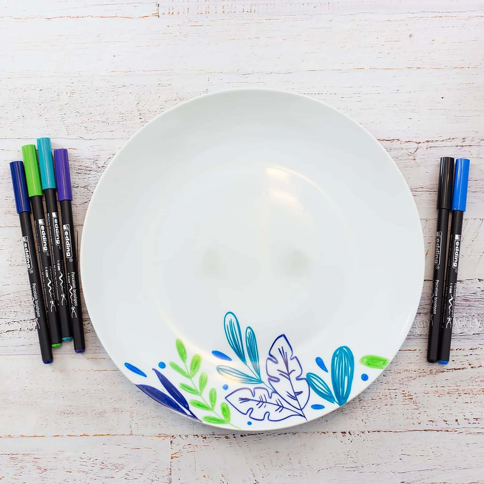 Ein weißer Teller mit blauen und grünen Blattmotiven, dekoriert mit dem edding 4200 Porzellanpinselstift von edding, ist von sechs farbigen Markern (blau, grün, schwarz) auf einer weißen Holzoberfläche umgeben. Ideal zum Dekorieren von Keramik und spülmaschinenfest.