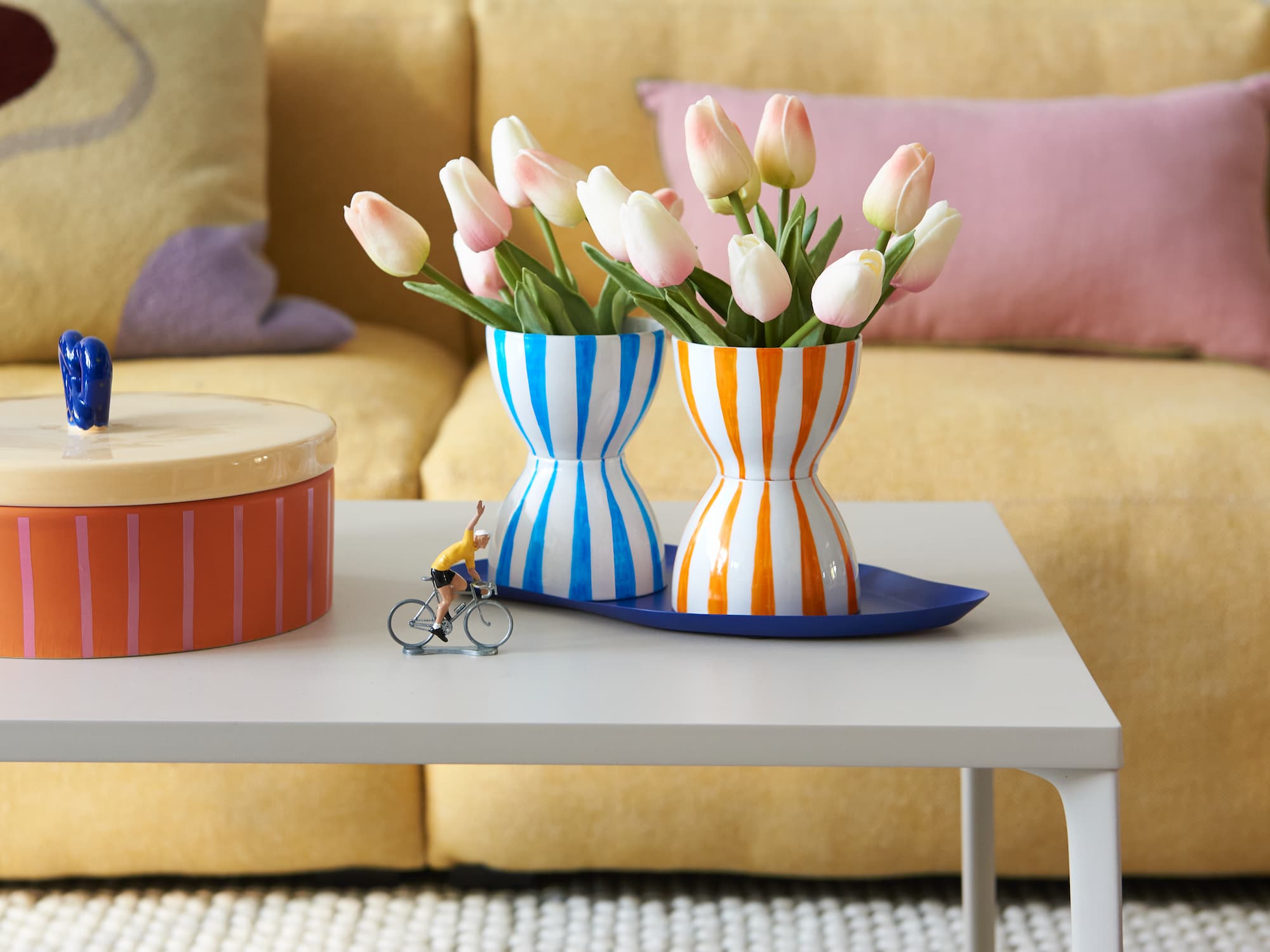 Auf einem weißen Couchtisch stehen zwei gestreifte Vasen mit rosa Tulpen, ein runder Behälter mit Deckel und eine kleine Radfahrerfigur – perfekt für Keramikdekoration mit dem edding 4200 Porzellanpinselstift von edding. Im Hintergrund steht ein gelbes Sofa mit bunten Kissen.
