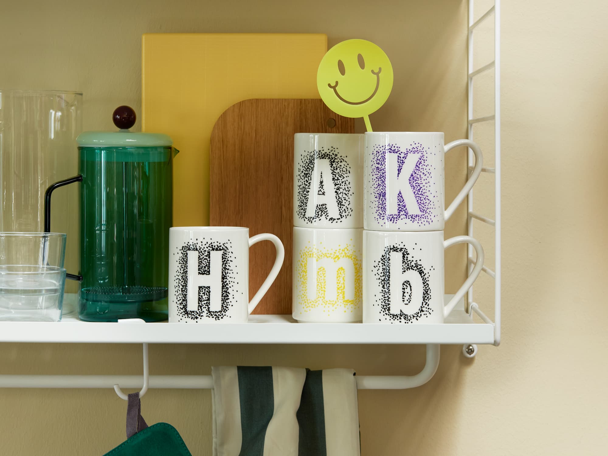 Vier weiße Tassen mit den schwarzen Buchstaben „H“, „A“, „b“ und dem gelben „m“, dekoriert mit dem edding 4200 Porzellanpinselstift von edding, stehen auf einem Regal neben einer grünen Kaffeepresse, Gläsern und einem Rührstab mit Smiley. Die Tassen sind spülmaschinenfest.