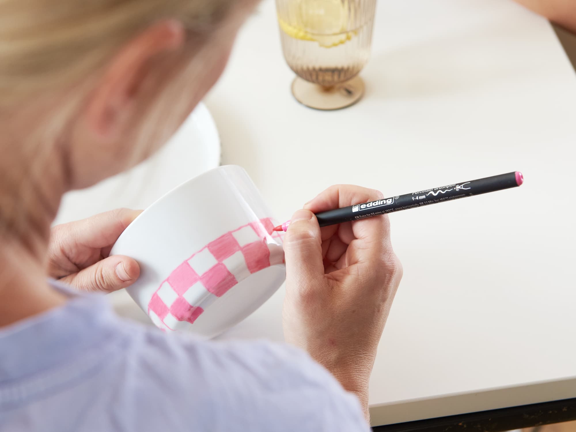 Mit dem edding 4200 Porzellanpinselstift von edding verziert eine Person eine weiße Tasse mit einem rosa Karomuster, während sie an einem mit einem Glas Wasser und einem Teller gedeckten Tisch sitzt.